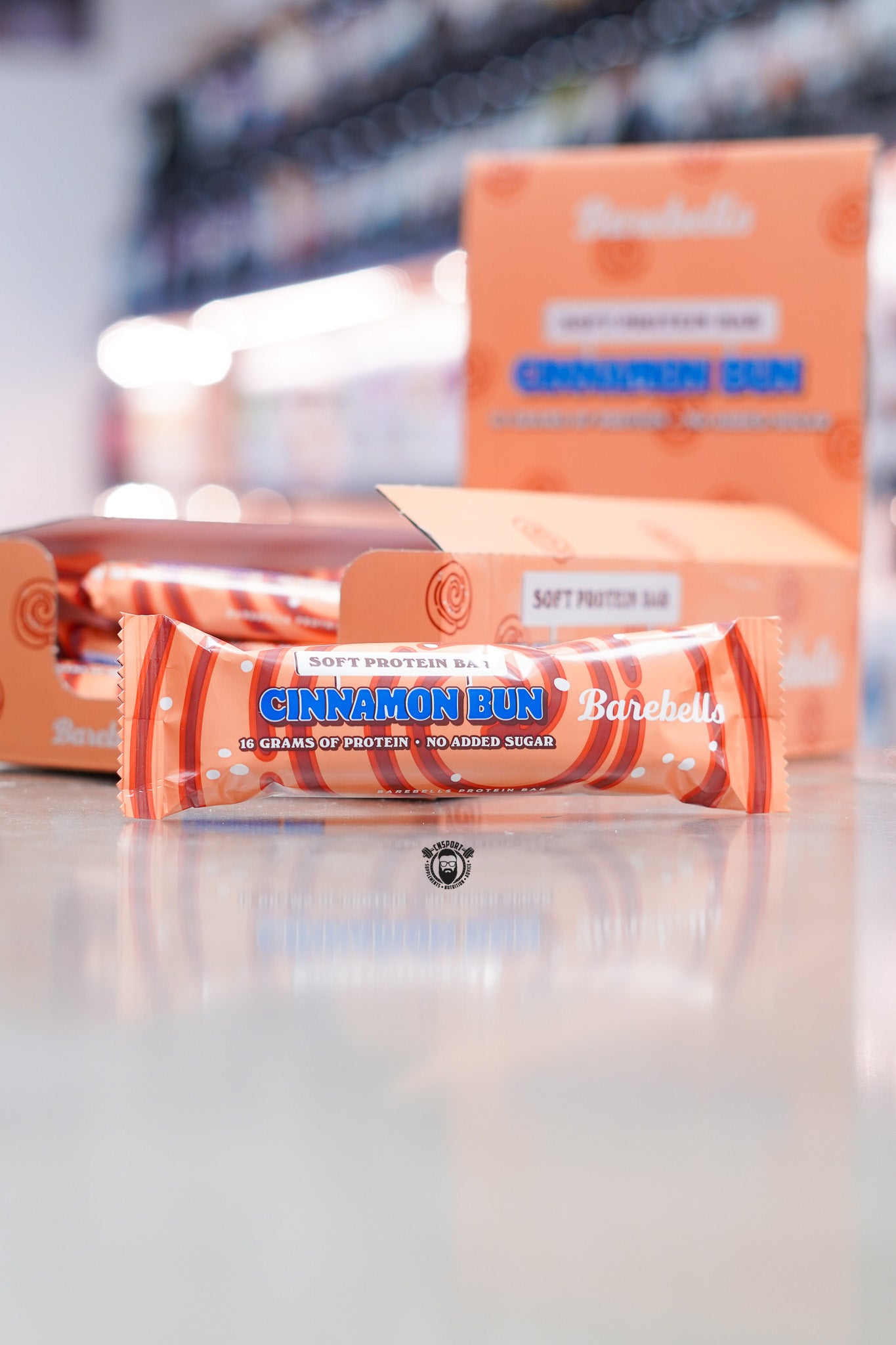 Barebells Soft Protein Bar Cinnamon Bun flavour with 16 grams of protein and no added sugar displayed in front of retail display boxes on supplement store counter with blurred background