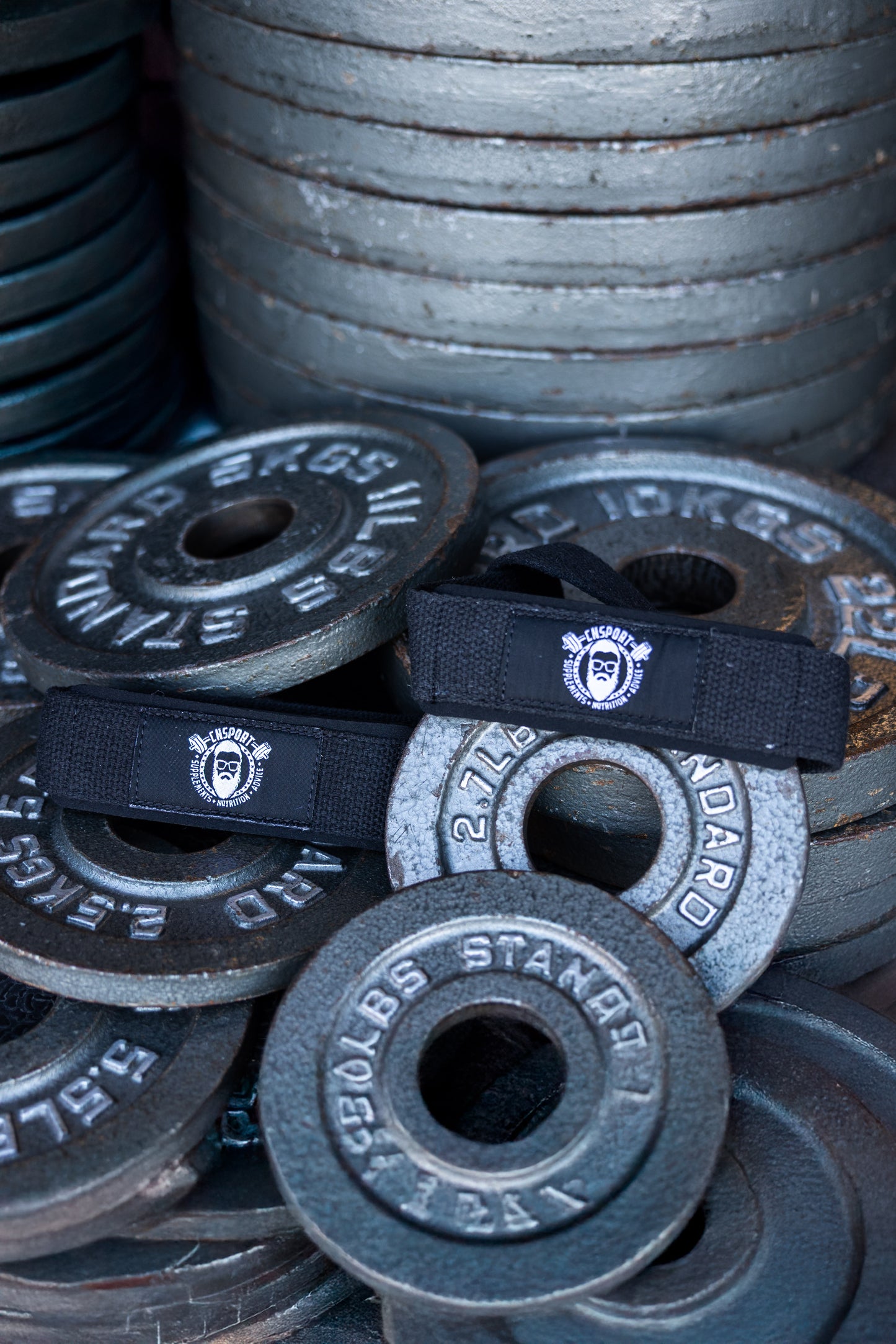 A pair of black CNSport cotton lifting straps with the CNSport logo placed on top of stacked metal weight plates in a gym setting.