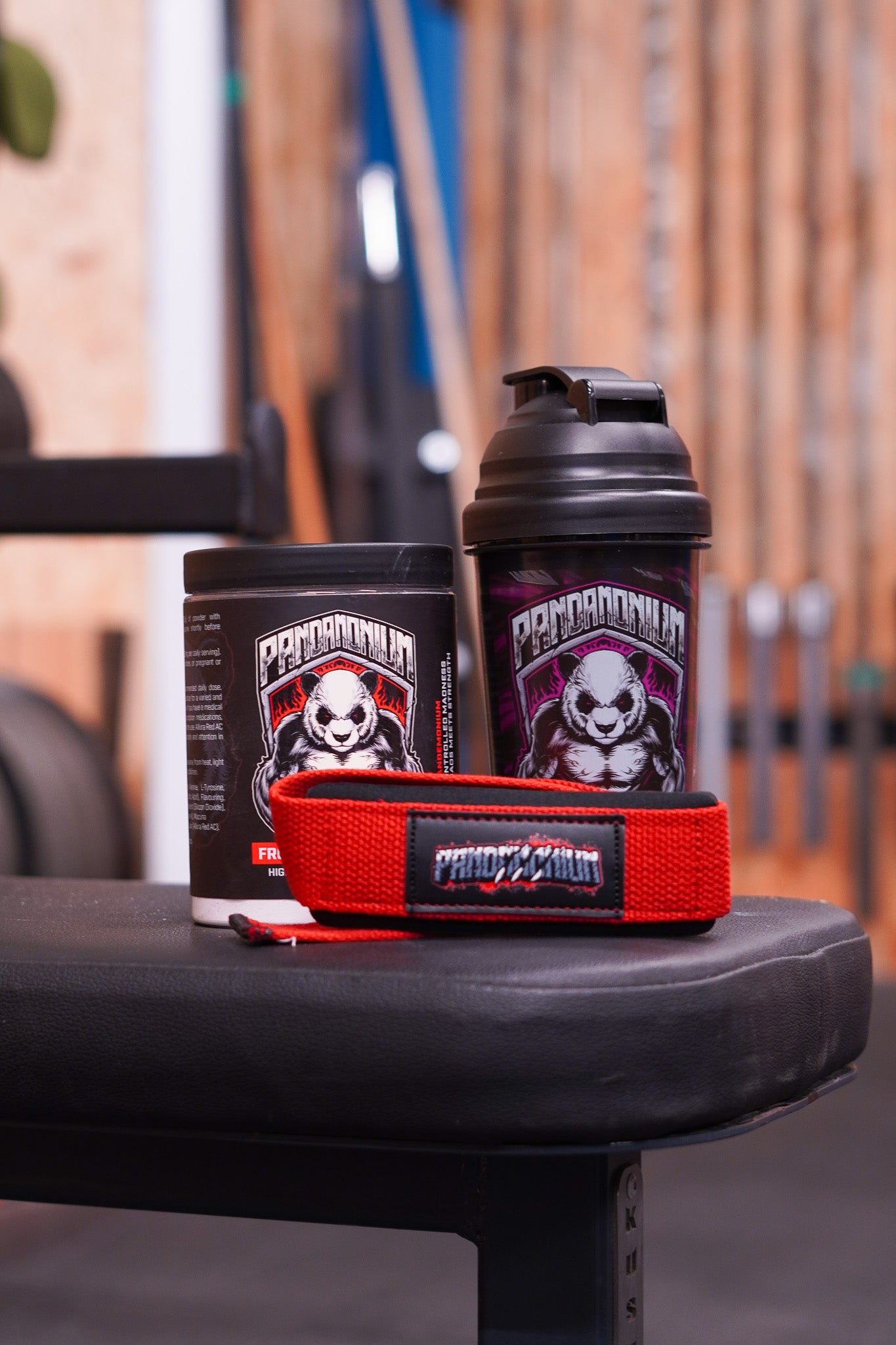 A gym bench display featuring a CNS Pandamonium Fruit Punch pre-workout tub and a black Pandamonium shaker bottle, with a pair of red Pandamonium lifting straps placed in front. Gym equipment is visible blurred in the background.