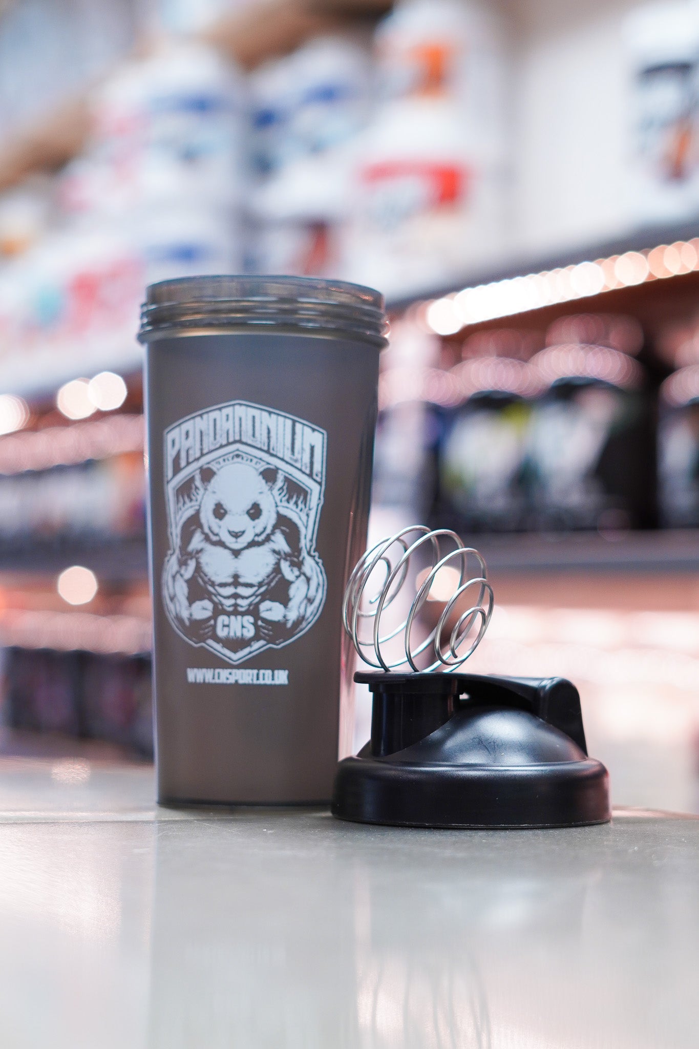 CNSport Pandamonium 600ml black shaker bottle with panda logo, shown with separate lid and metal mixing ball on a counter in a supplement store.