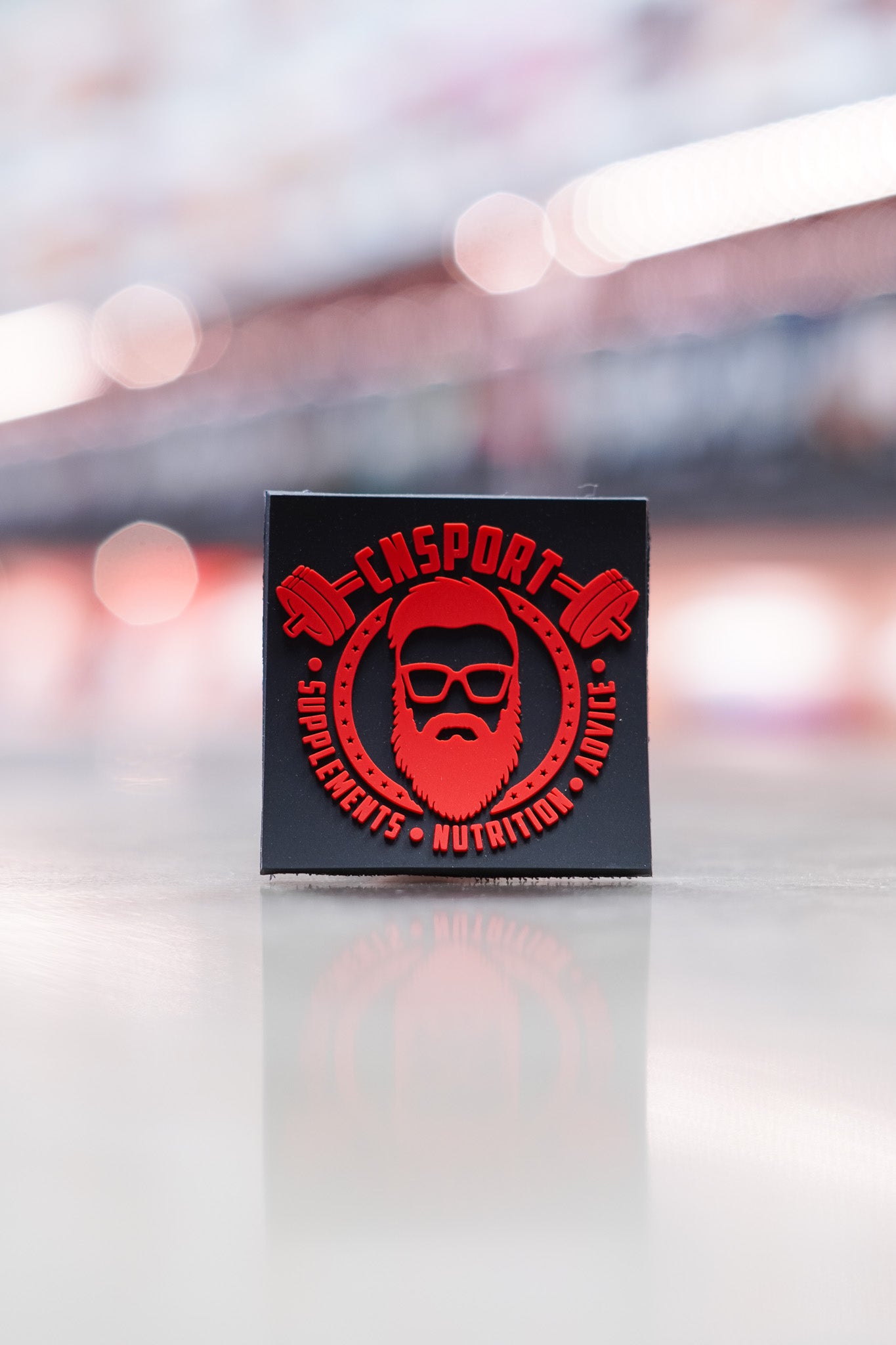 Black CNSport velcro patch with striking red embossed branding and barbell artwork, reading “Supplements • Nutrition • Advice,” shown in a retail setting with a soft bokeh background.