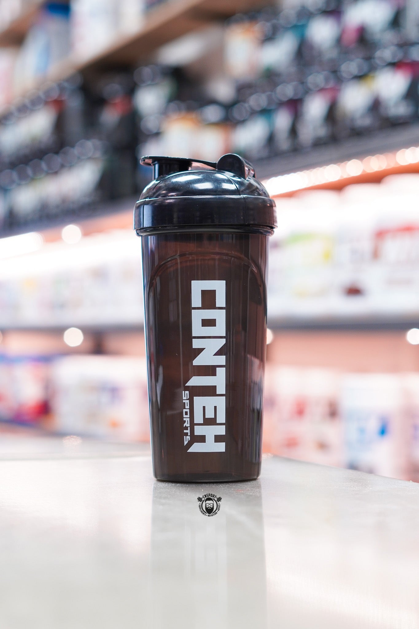 Smoky black Conteh Sports 700ml ball shaker bottle with black flip-top lid displayed on a supplement shop counter.