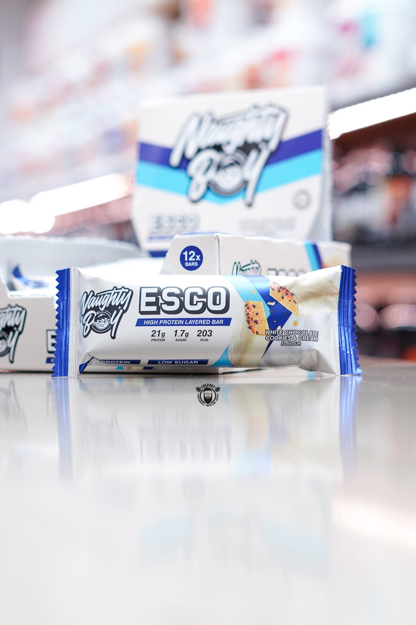 Naughty Boy ESCO high protein layered bar, White Chocolate Cookies & Cream flavour (21g protein), displayed in front of 12-bar retail box in supplement store.