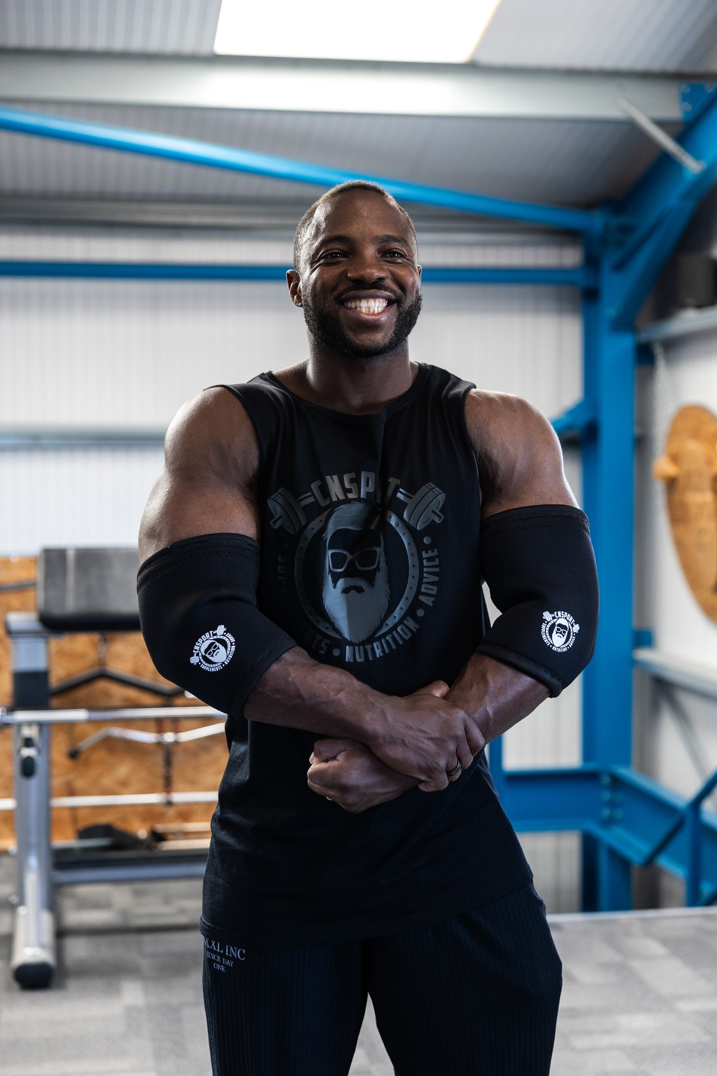 A muscular man in a black CNSport tank top adjusts a black elbow sleeve branded with the CNSport logo while standing in a gym with dumbbells in the background.
