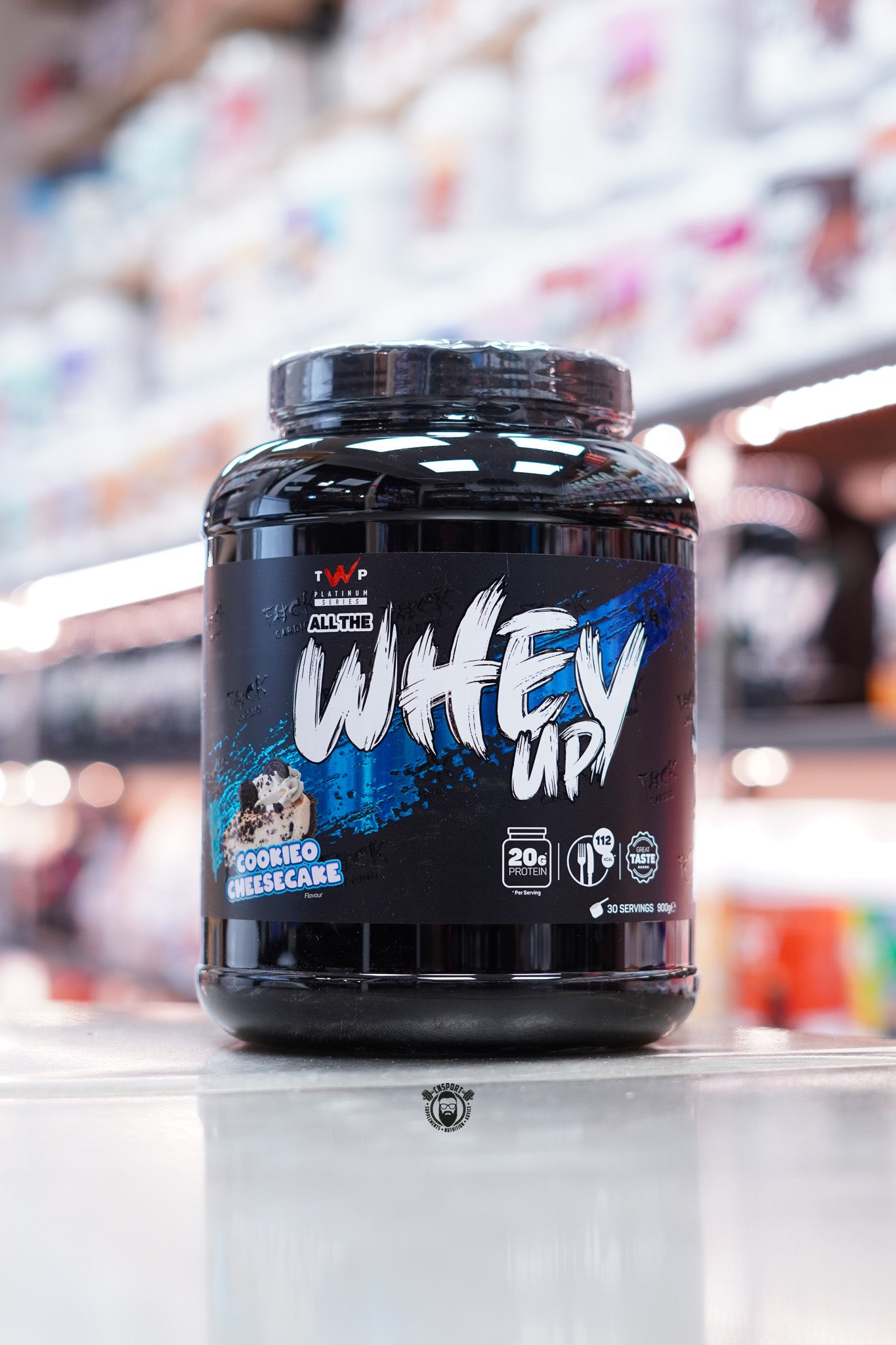 900g tub of TWP All The Whey Up protein powder in Cookieo Cheesecake flavour, featuring bold white and blue branding with a dessert image. The label highlights 20g protein, 112 calories per serving, and includes “Great Taste” and 30 servings icons.