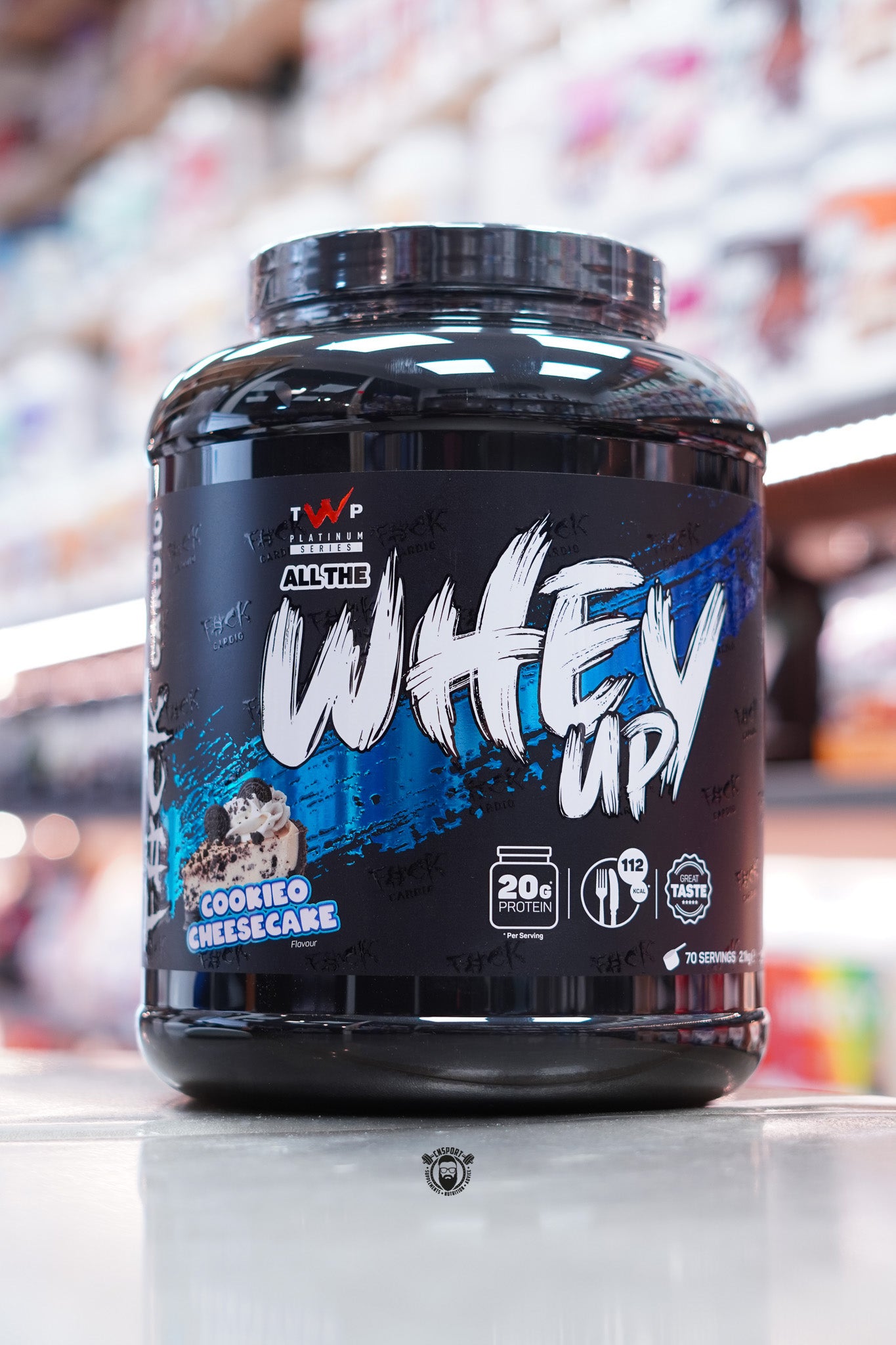 Large black tub of TWP All The Whey Up protein powder in Cookieo Cheesecake flavour, featuring bold white and blue branding with an image of a cheesecake topped with cookies. Label highlights 20g protein and 112 calories per serving.
