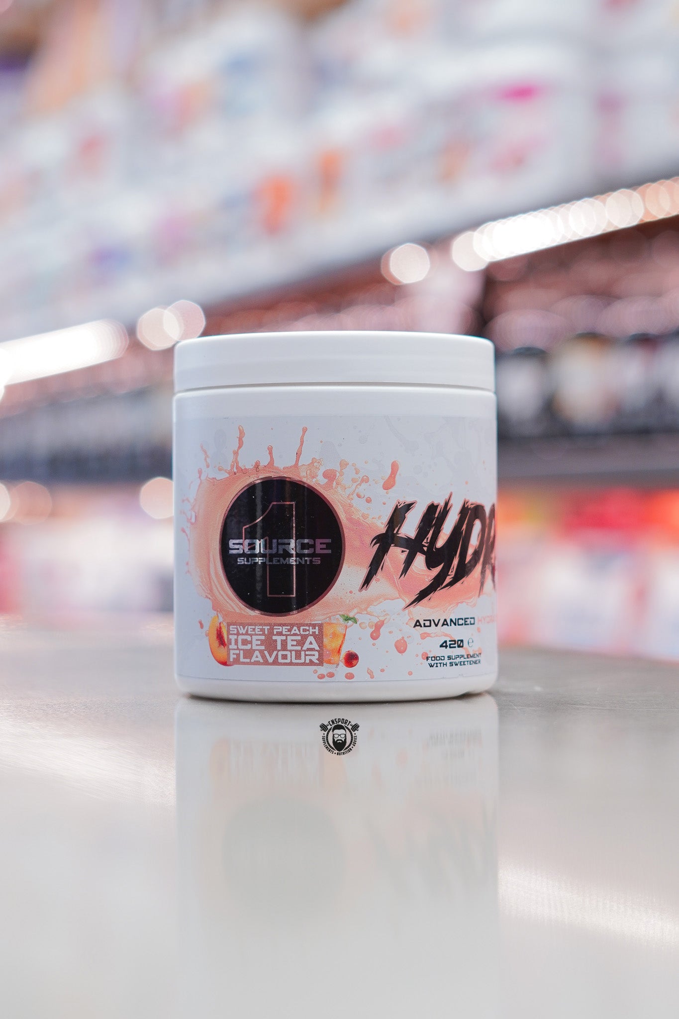 1 Source Supplements Hydrate advanced hydration powder tub in Sweet Peach Ice Tea flavour displayed on a retail counter with blurred supplement shelves behind.