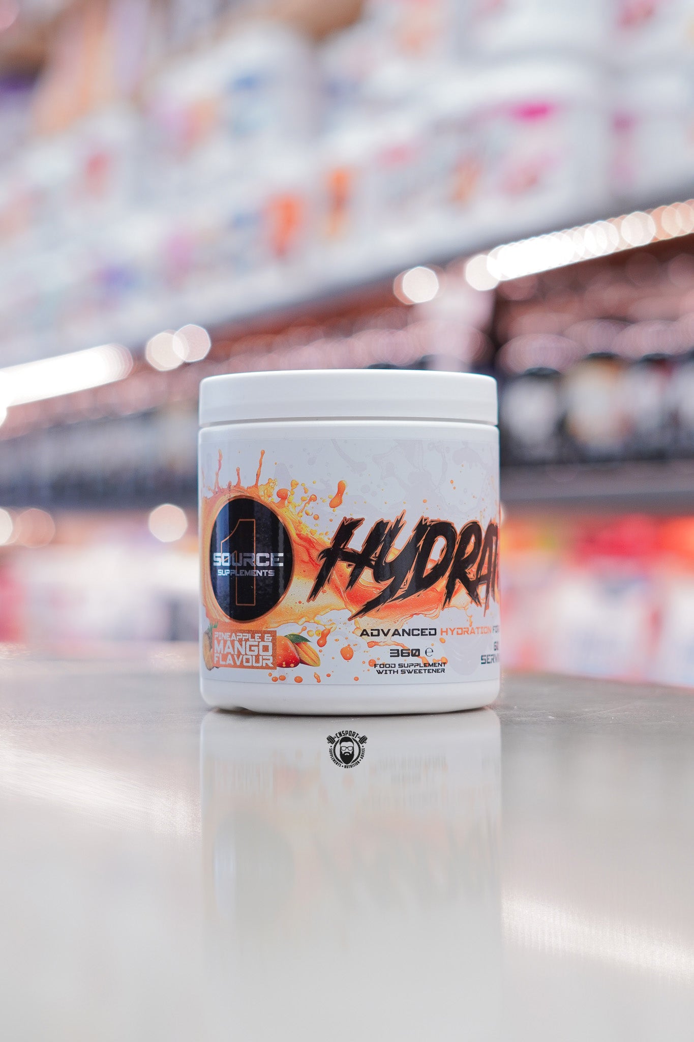 1 Source Supplements Hydrate advanced hydration powder tub in Pineapple & Mango flavour on a retail counter with blurred supplement shelves in the background.