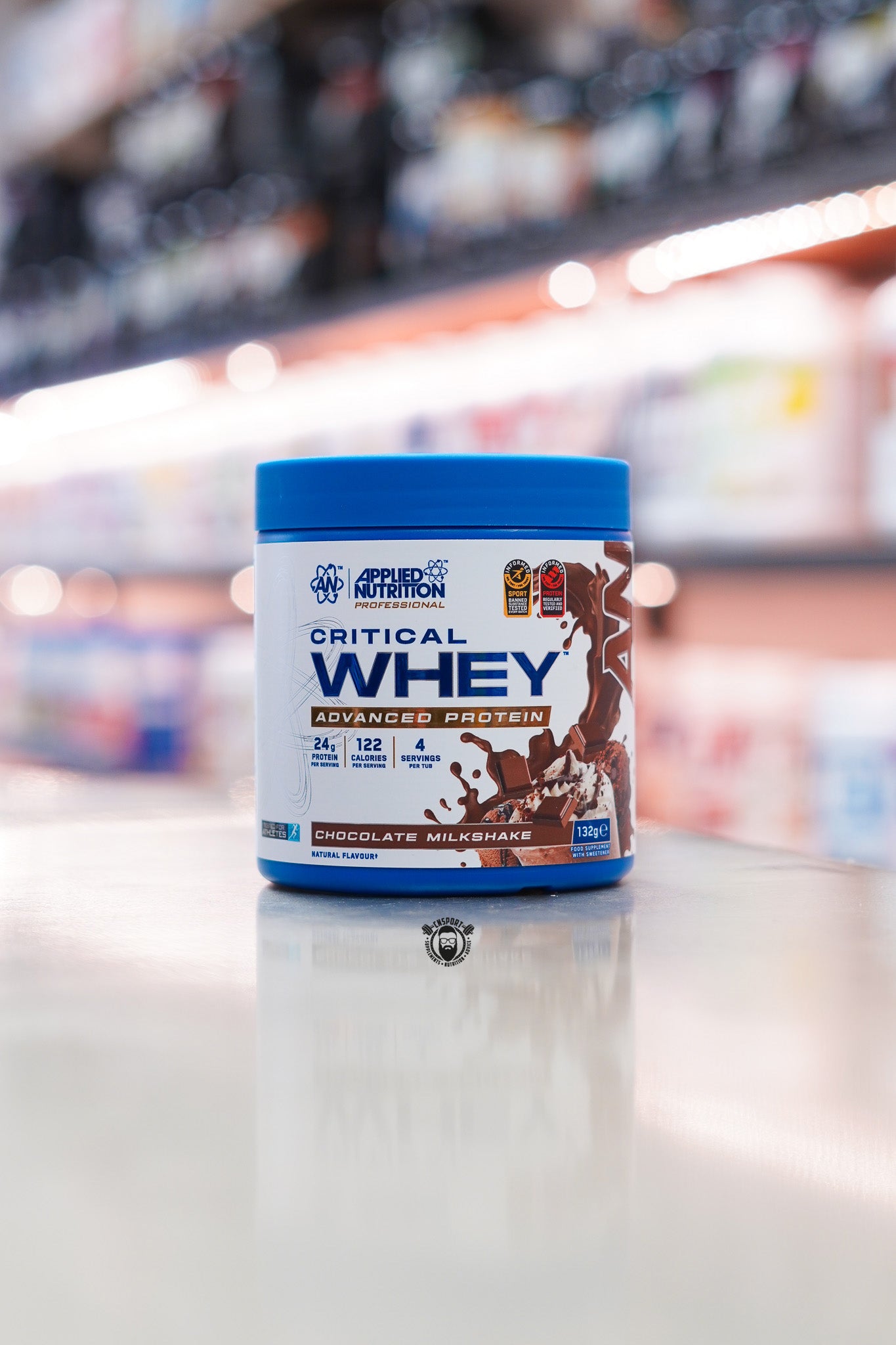 Applied Nutrition Critical Whey 132g protein powder sample tub, Chocolate Milkshake flavour, displayed on a supplement store counter.
