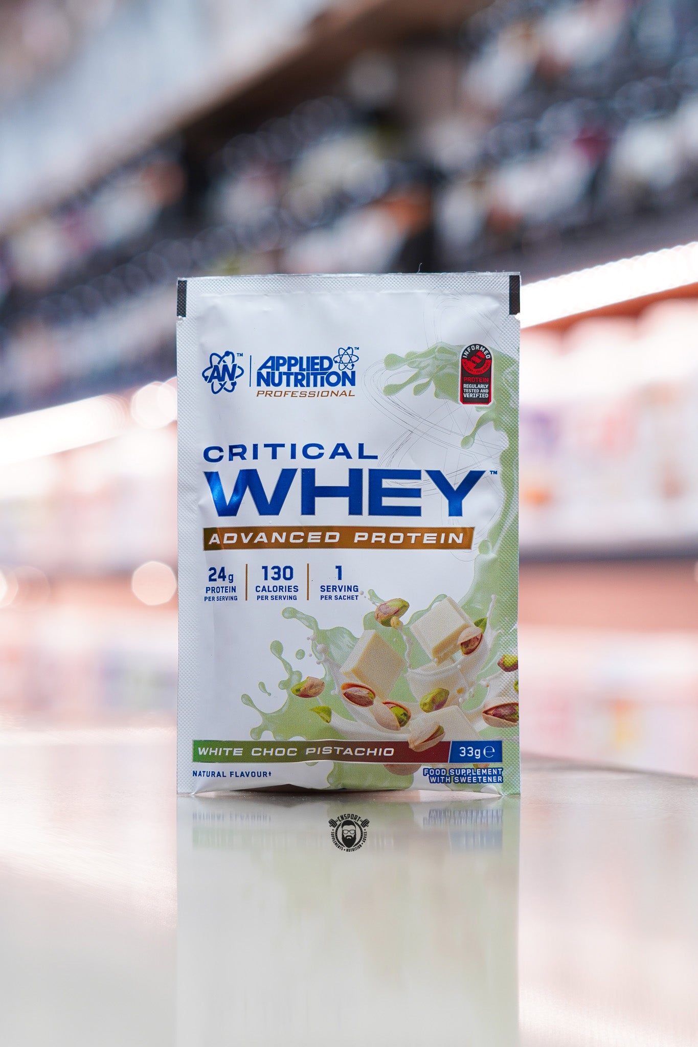 Applied Nutrition Critical Whey Advanced Protein sample sachet in White Choc Pistachio flavour displayed on a supplement store counter, featuring 24g protein per serving with white chocolate and pistachio imagery.