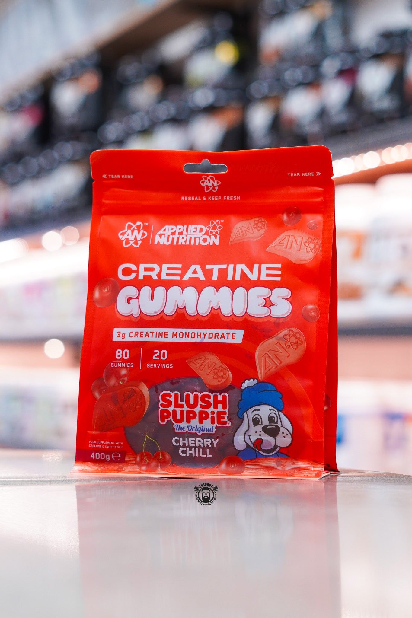 Applied Nutrition Creatine Gummies Cherry Chill pouch displayed on supplement store counter with blurred retail shelving background, featuring 3g creatine monohydrate per serving, 80 gummies, 20 servings, and Slush Puppie Cherry Chill flavour branding.