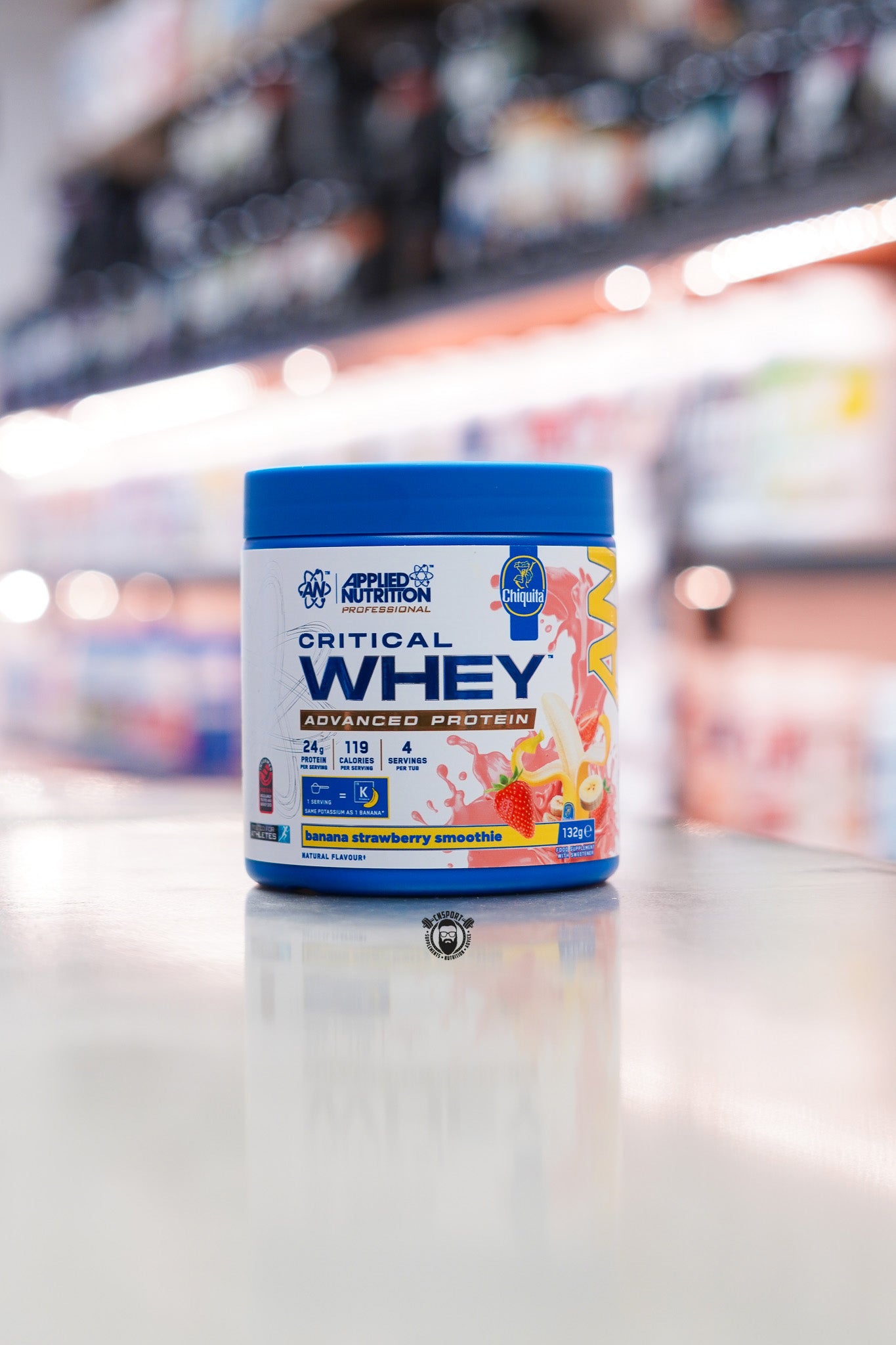 Applied Nutrition Critical Whey 132g protein powder sample tub, Banana Strawberry Smoothie flavour, displayed on a supplement store counter.