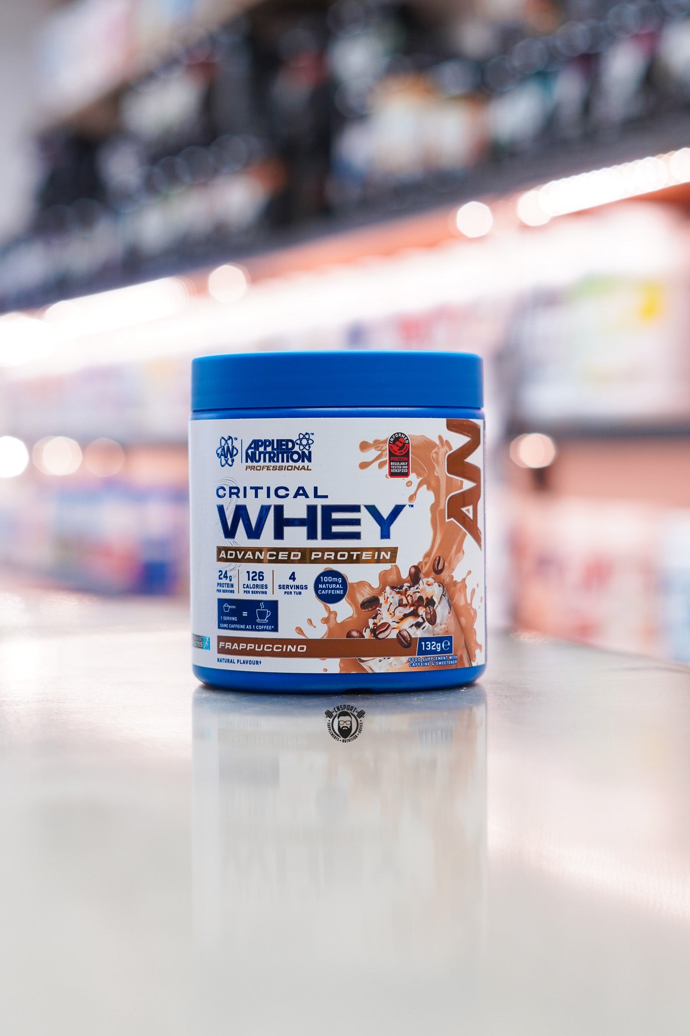 Applied Nutrition Critical Whey 132g protein powder sample tub, Frappuccino flavour, displayed on a supplement store counter.