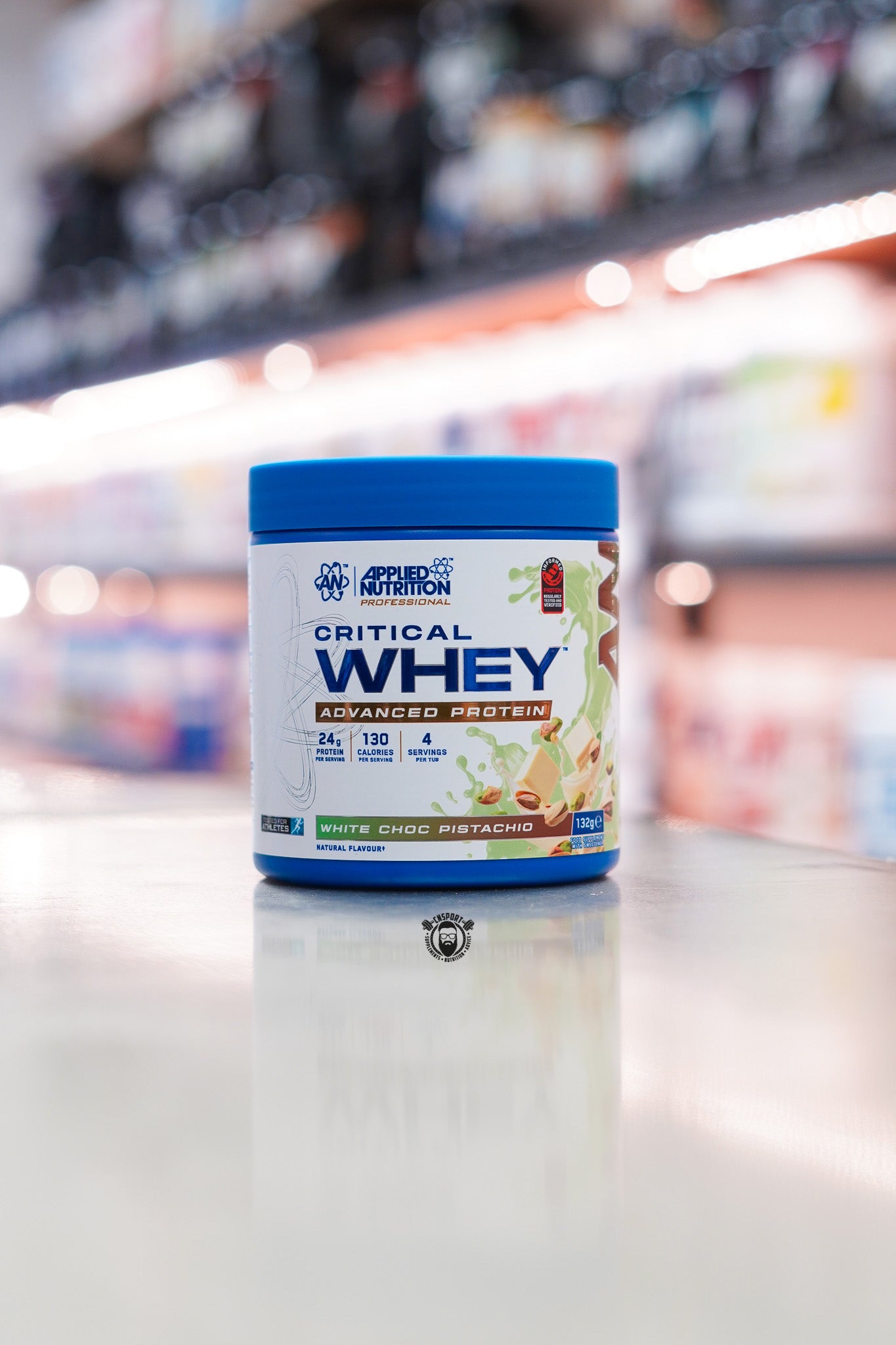 Applied Nutrition Critical Whey 132g protein powder sample tub, White Choc Pistachio flavour, displayed on a supplement store counter.