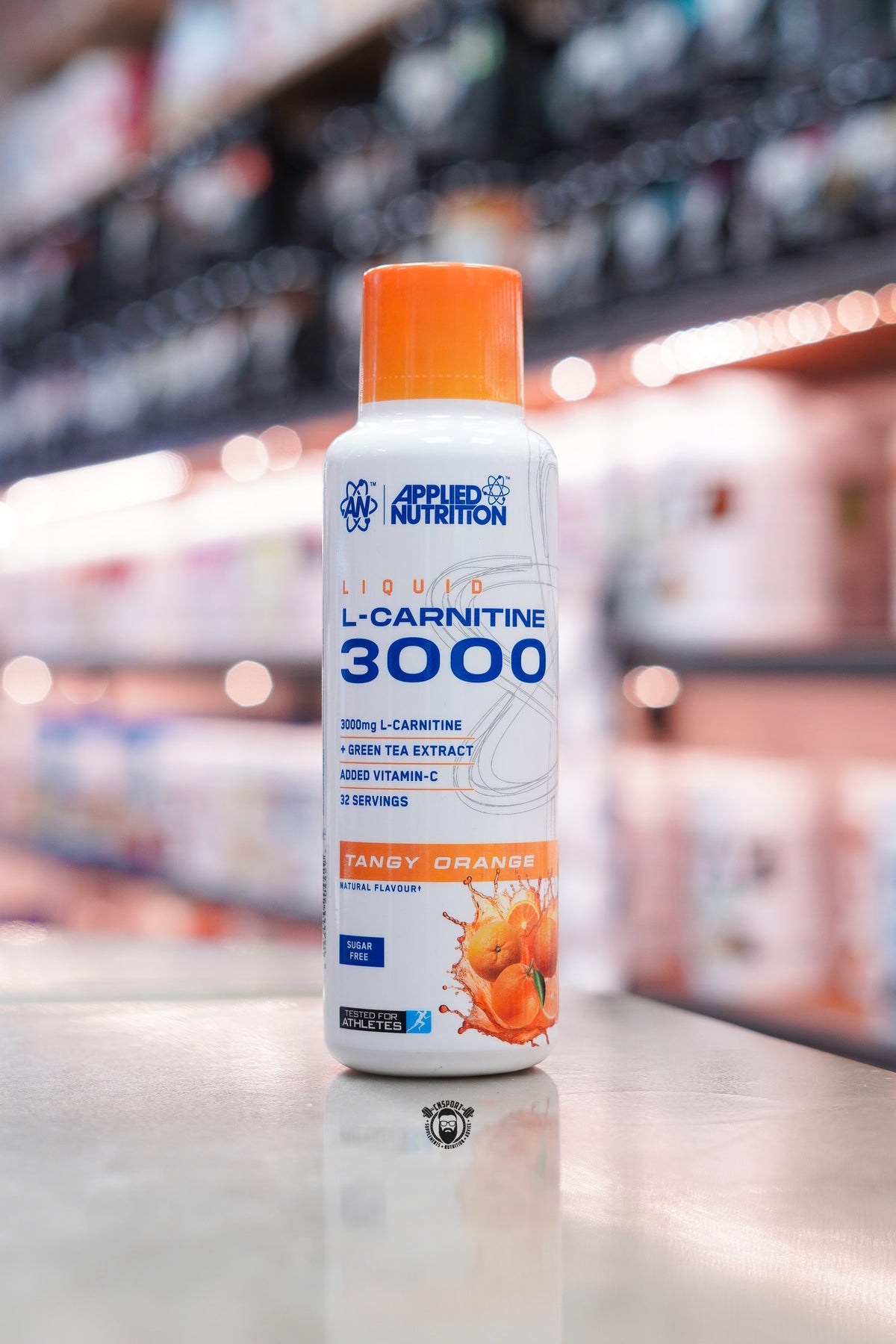 Applied Nutrition L-Carnitine Liquid 3000 Tangy Orange flavour bottle, sugar free with 32 servings, displayed on a supplement shop counter.