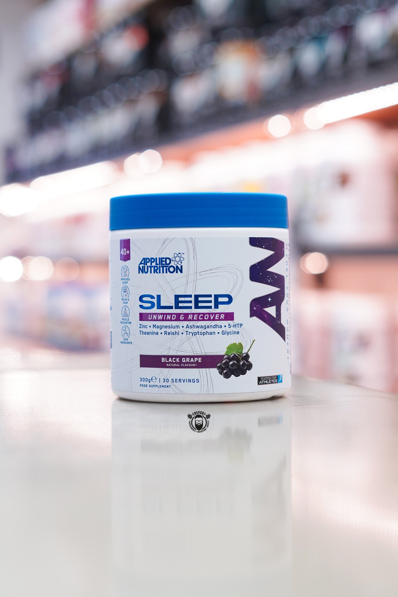 Applied Nutrition Sleep Unwind & Recover in Black Grape flavour, 300g tub with 30 servings, displayed on a countertop in a supplement store.