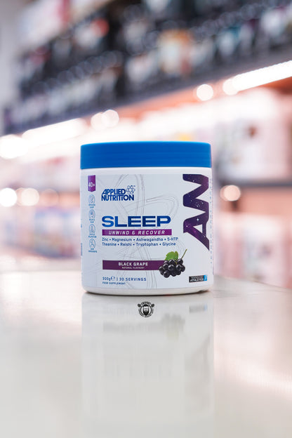 Applied Nutrition Sleep Unwind & Recover in Black Grape flavour, 300g tub with 30 servings, displayed on a countertop in a supplement store.