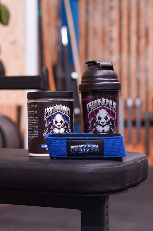 A gym bench display featuring a CNS Pandamonium Blue Raspberry pre-workout tub and a black Pandamonium shaker bottle, with a pair of blue Pandamonium lifting straps positioned in front. The background shows gym equipment with soft blur.