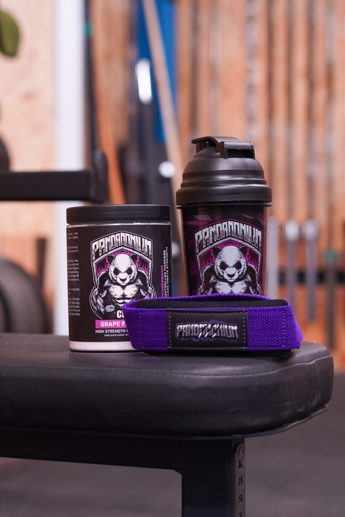 A gym bench display featuring a CNS Pandamonium Grape pre-workout tub and a black Pandamonium shaker bottle, with a pair of purple Pandamonium lifting straps sitting in front. The background shows out-of-focus gym equipment.