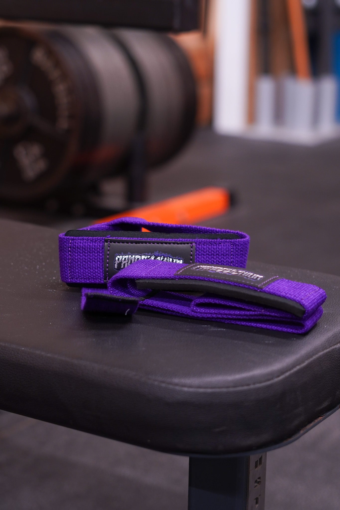 Purple Pandamonium lifting straps placed on a gym bench with weight plates softly blurred in the background.