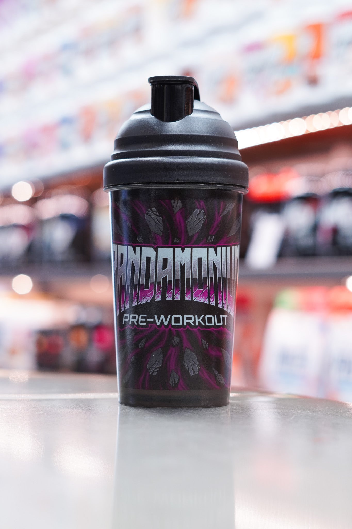 Black CNSport Pandamonium 600ml shaker standing on a reflective surface, featuring a bold purple and grey cracked-stone design with the “PANDAMONIUM PRE-WORKOUT” logo on the front. Gym supplement shelves are blurred in the background.