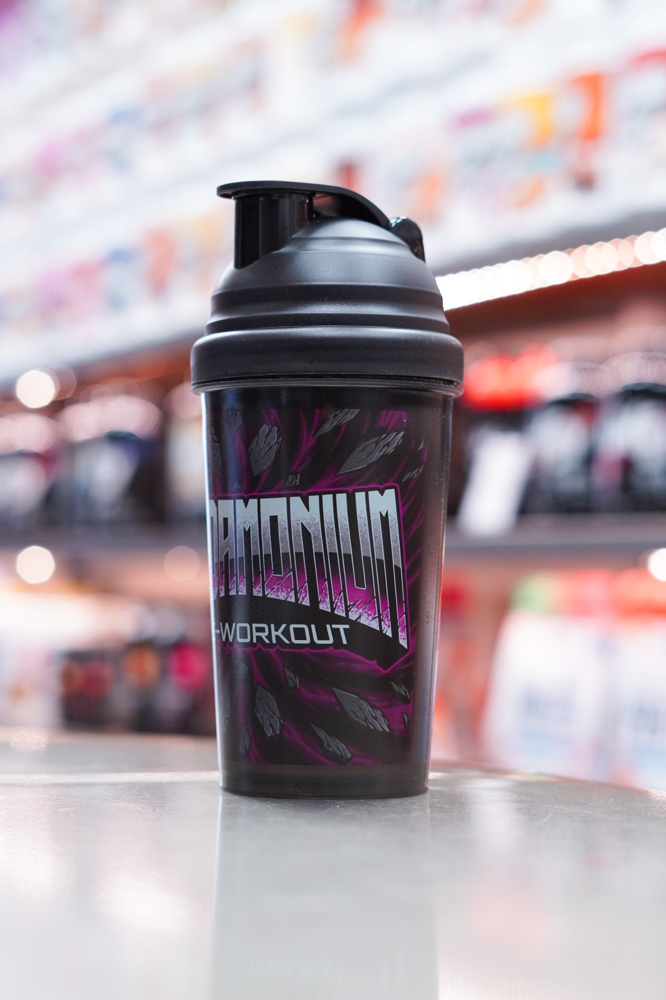 Side-angled view of the black CNSport Pandamonium shaker showing the stylised “PANDAMONIUM PRE-WORKOUT” branding with purple graphic accents, photographed in a supplement store environment.