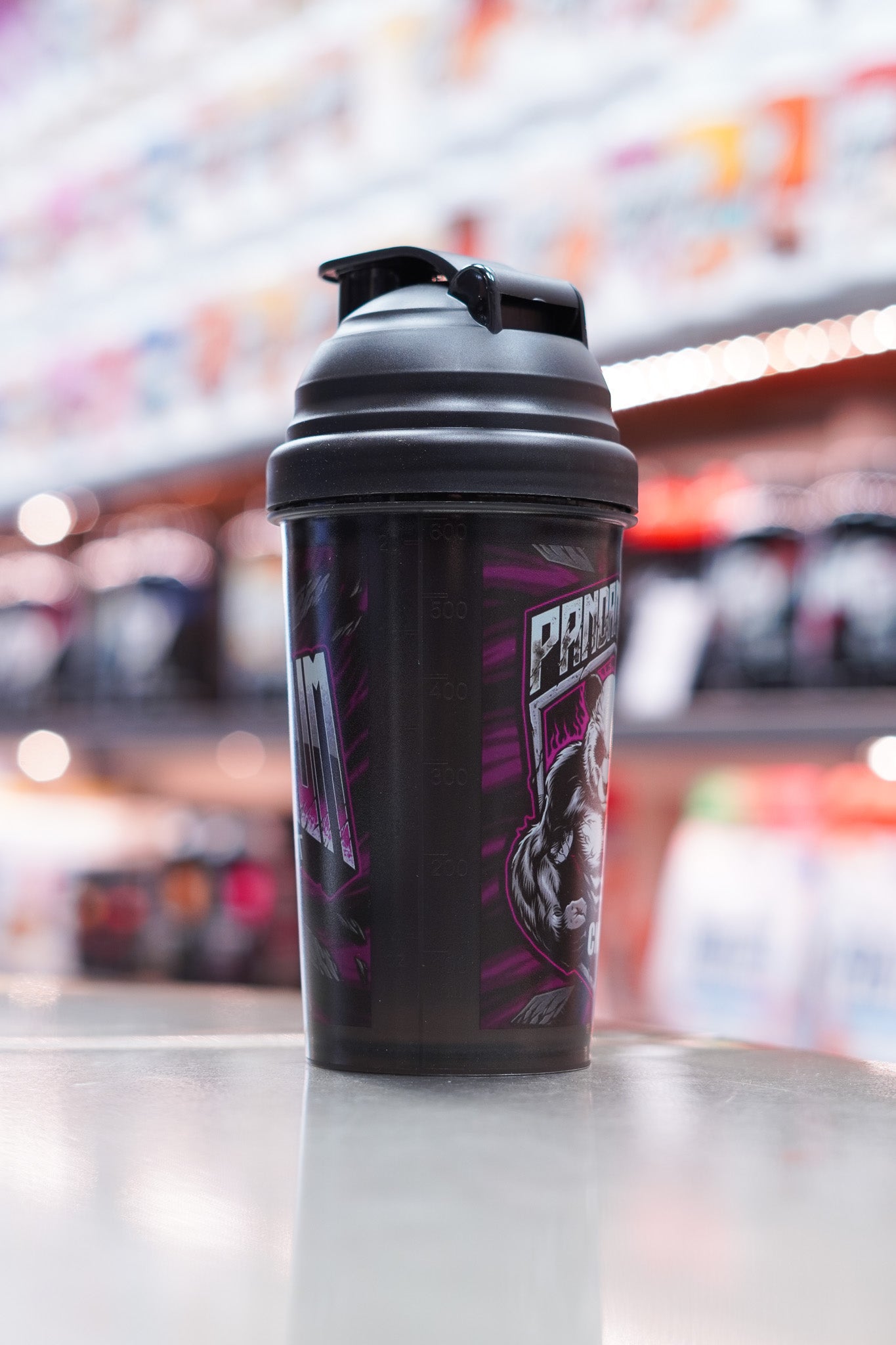 Side view of the shaker showing the measurement markings on the semi-transparent black body and part of the panda mascot graphic, with supplement shelves out of focus in the background.