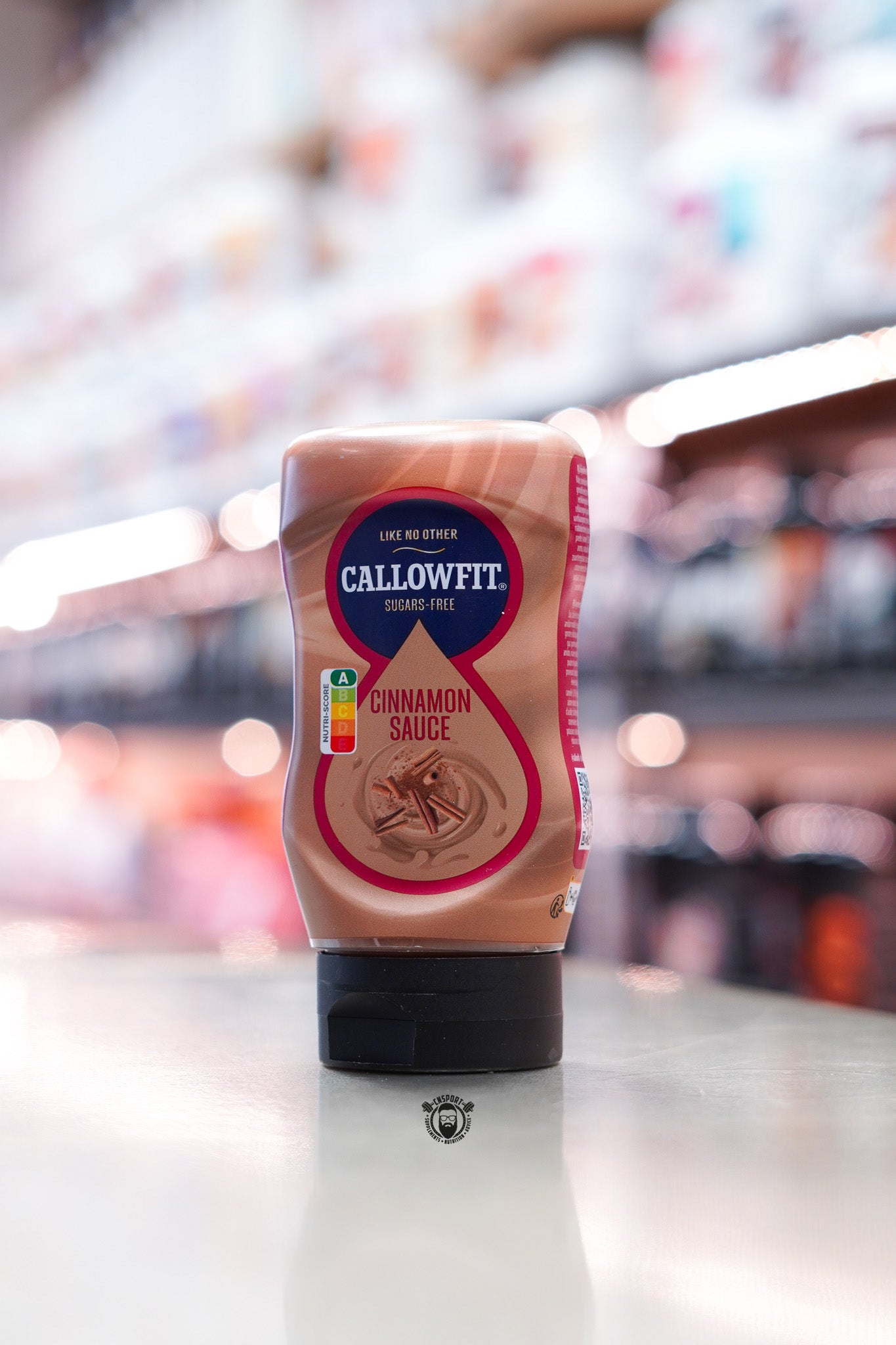 Callowfit Cinnamon Sauce 300ml sugar-free sauce bottle displayed upright with cinnamon flavour branding against a blurred retail supplement store background.
