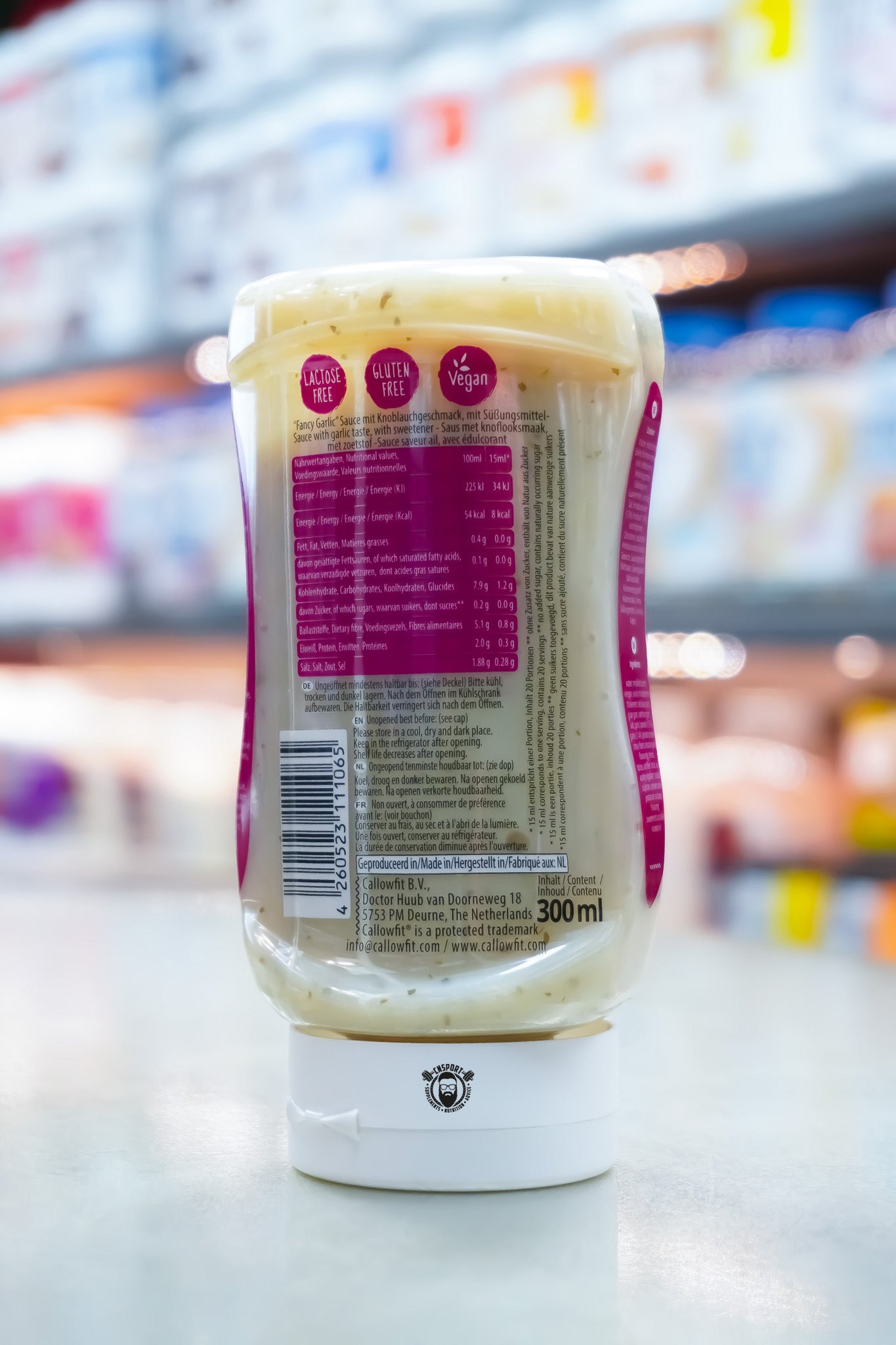 Rear view of Callowfit Fancy Garlic Sauce 300ml bottle displaying nutritional information, ingredients, barcode and storage instructions with a blurred supplement store background.