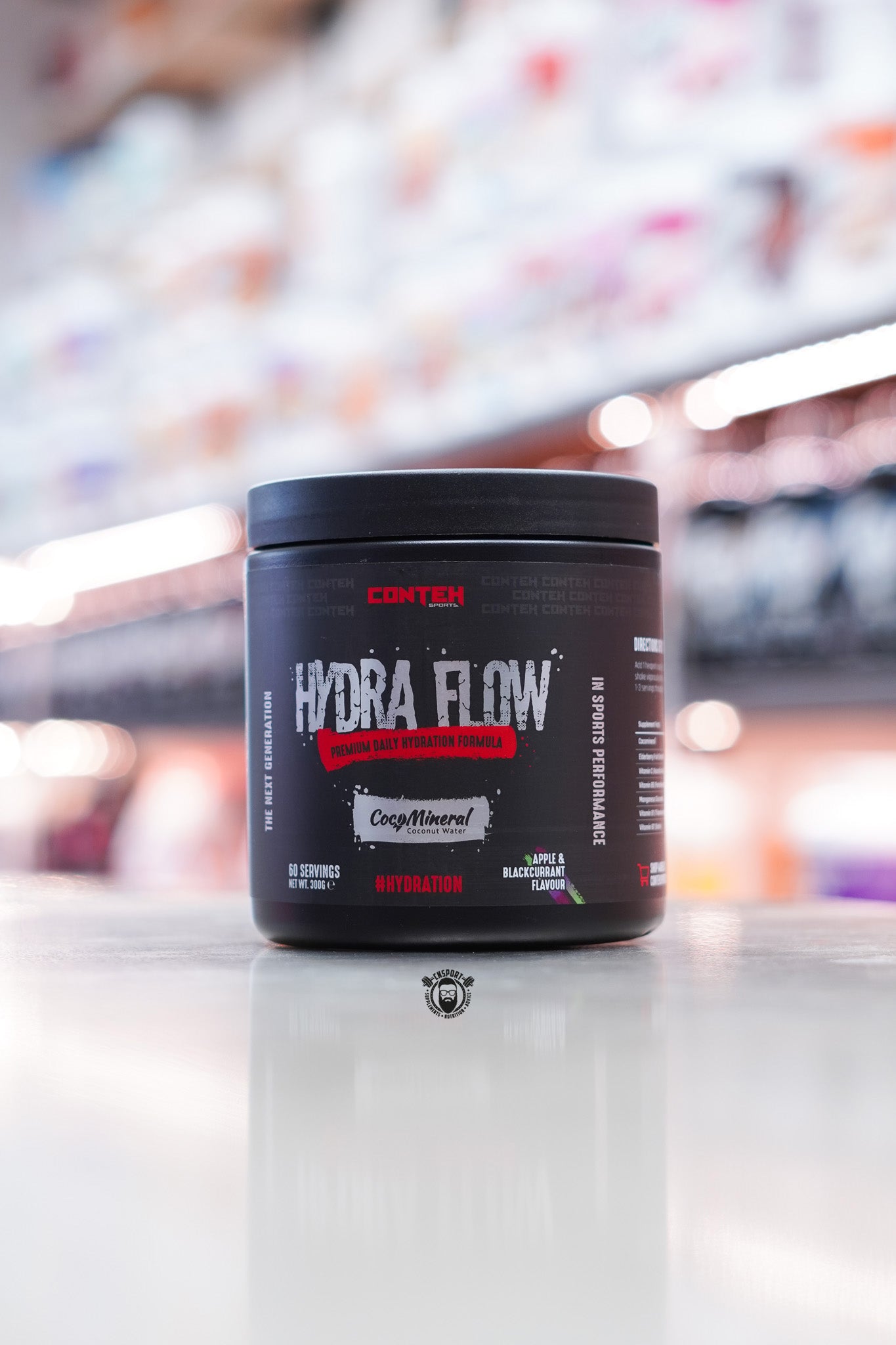 Conteh Sports Hydra Flow hydration supplement in Apple & Blackcurrant flavour – 60 servings with CocoMineral coconut water, black tub on shop counter.
