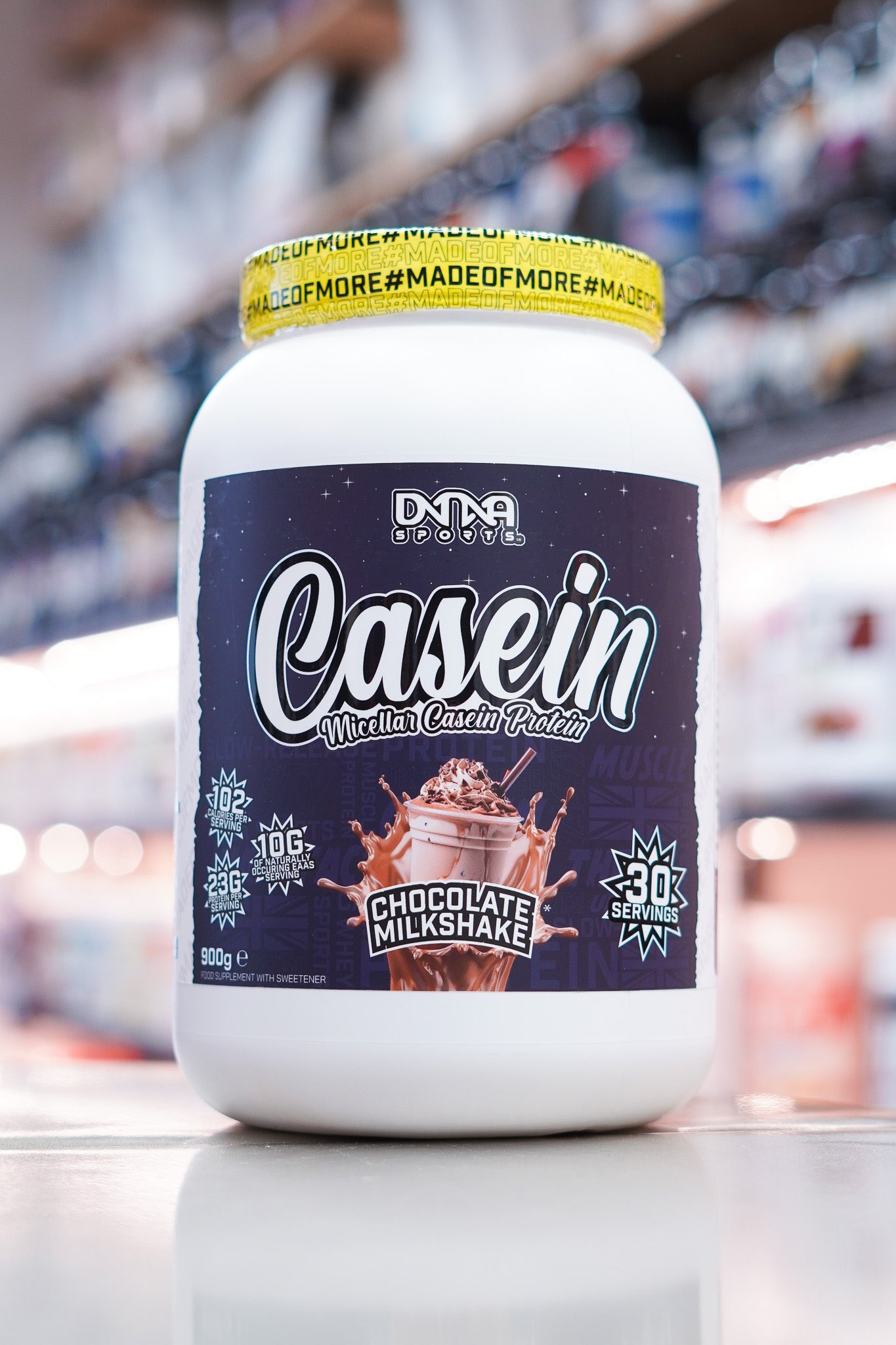 DNA Sports Casein Micellar Casein Protein Chocolate Milkshake 900g 30 servings tub on retail counter with blurred supplement store background