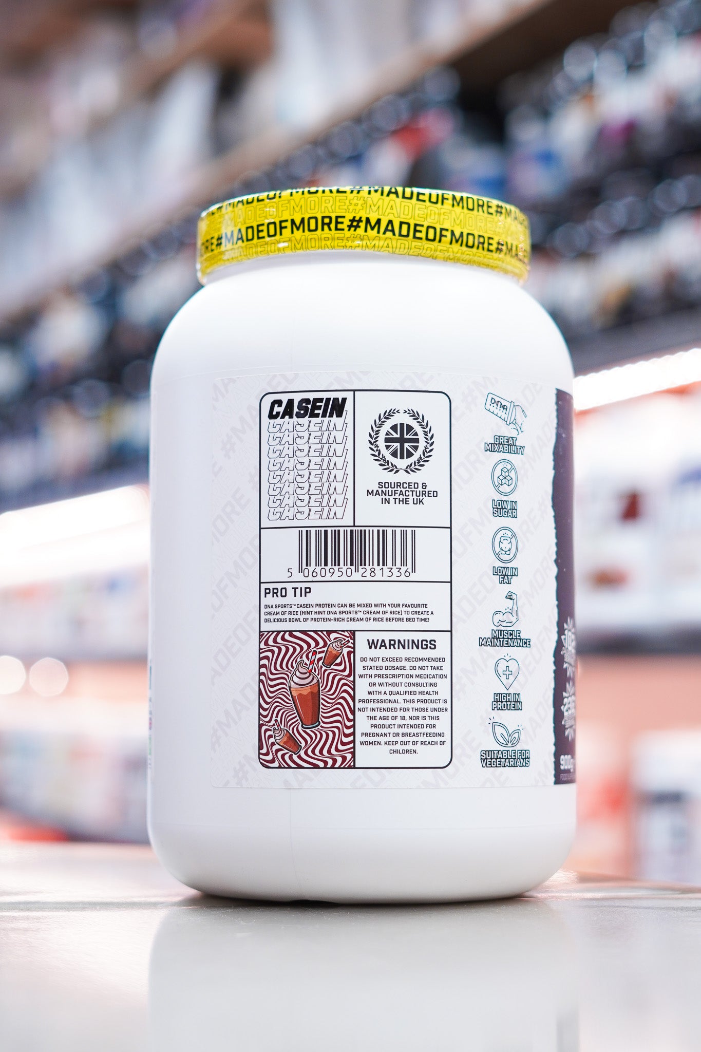DNA Sports Casein Micellar Casein Protein Chocolate Milkshake 900g side label showing product information and icons on retail counter with blurred supplement store background