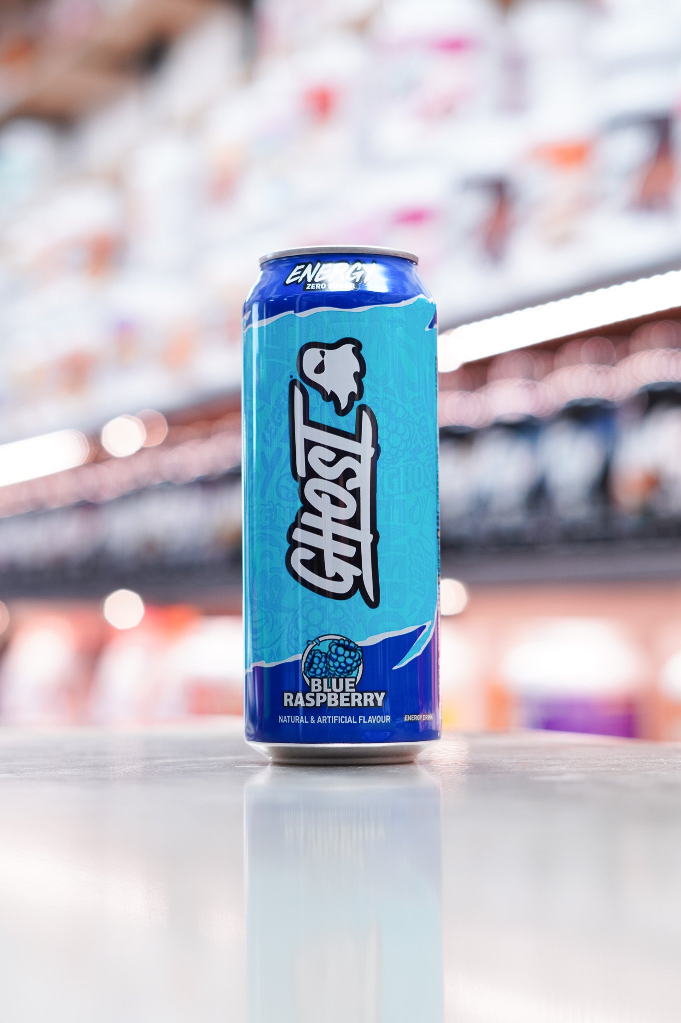 Tall can of Ghost Energy Drink in Blue Raspberry flavor, featuring a bold blue design with ghost logo, placed on a reflective surface in a well-lit supplement store.