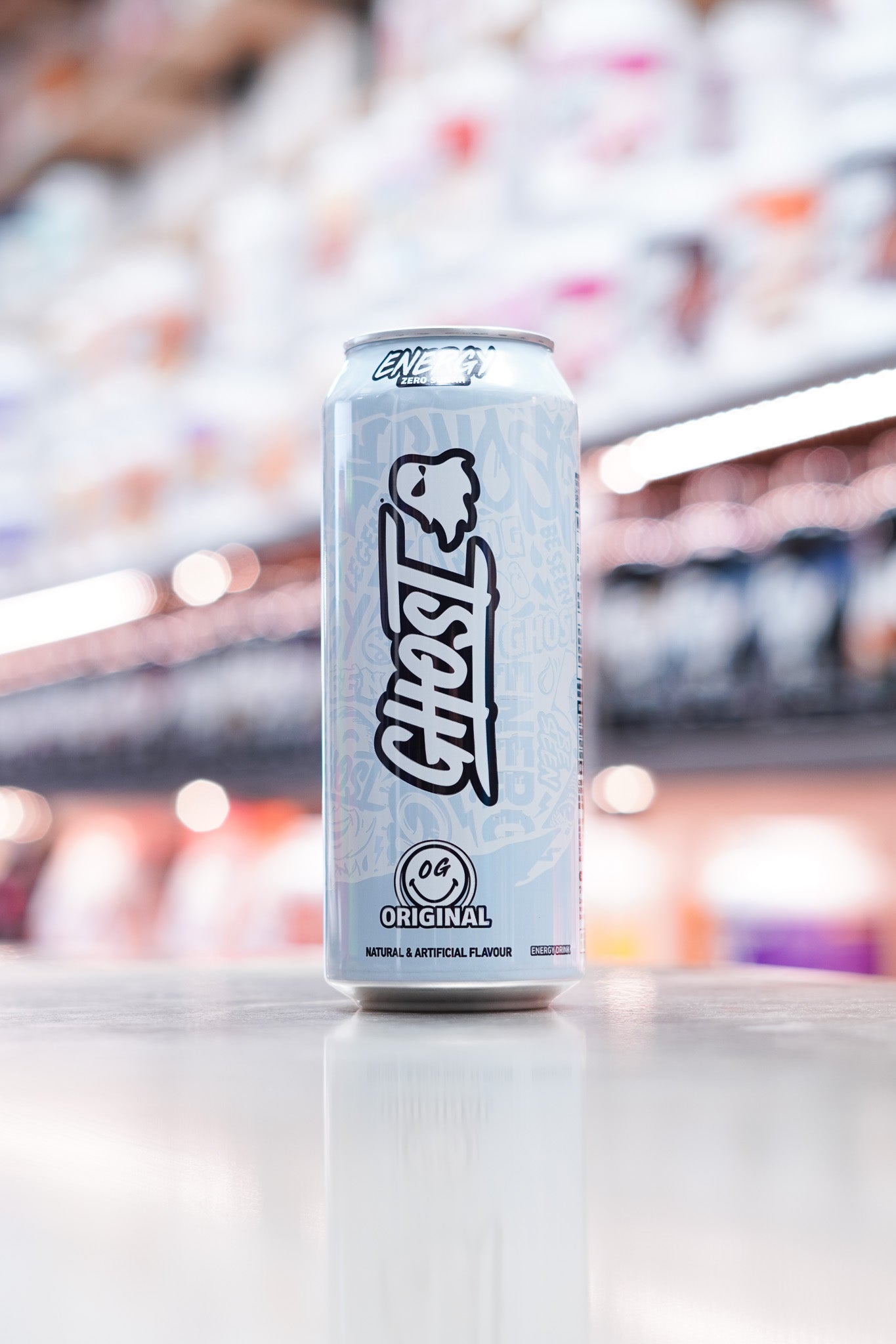 Minimalist white and grey Ghost Energy can labeled “Original”, standing upright on a reflective surface in front of shelves of colorful supplements.