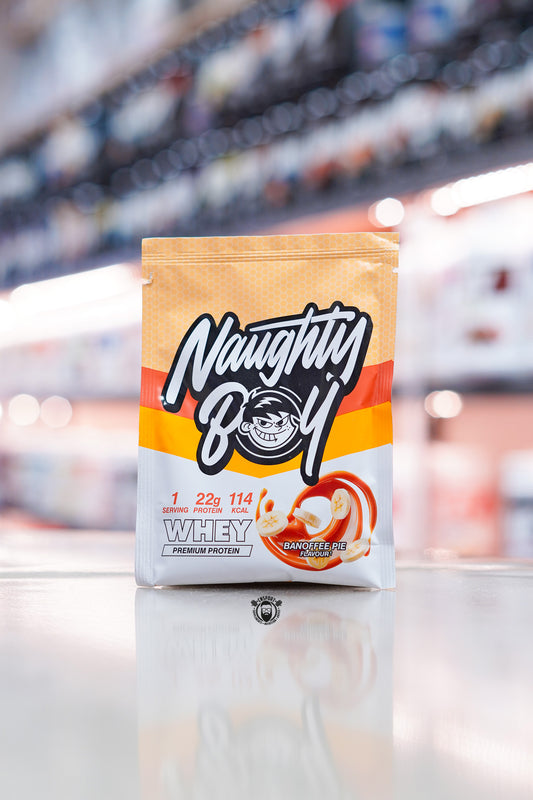 Naughty Boy Whey Protein 30g sample sachet Banoffee Pie flavour on retail counter with blurred supplement store background