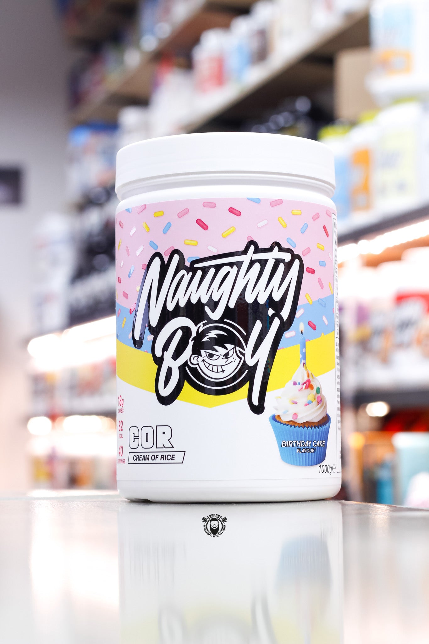 1kg tub of Naughty Boy COR (Cream of Rice) in Birthday Cake flavour, featuring a colorful sprinkle design and cupcake image on the label.