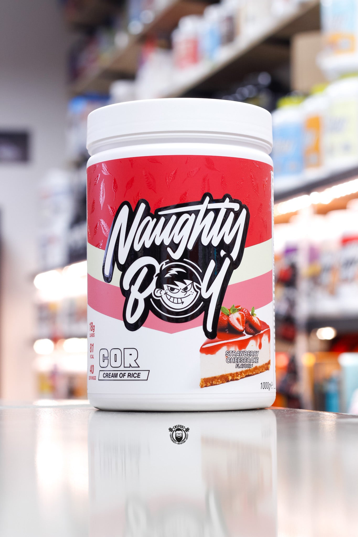 1kg tub of Naughty Boy COR Cream of Rice in Strawberry Cheesecake flavour, decorated with a red and pink label and a slice of cheesecake topped with strawberries.