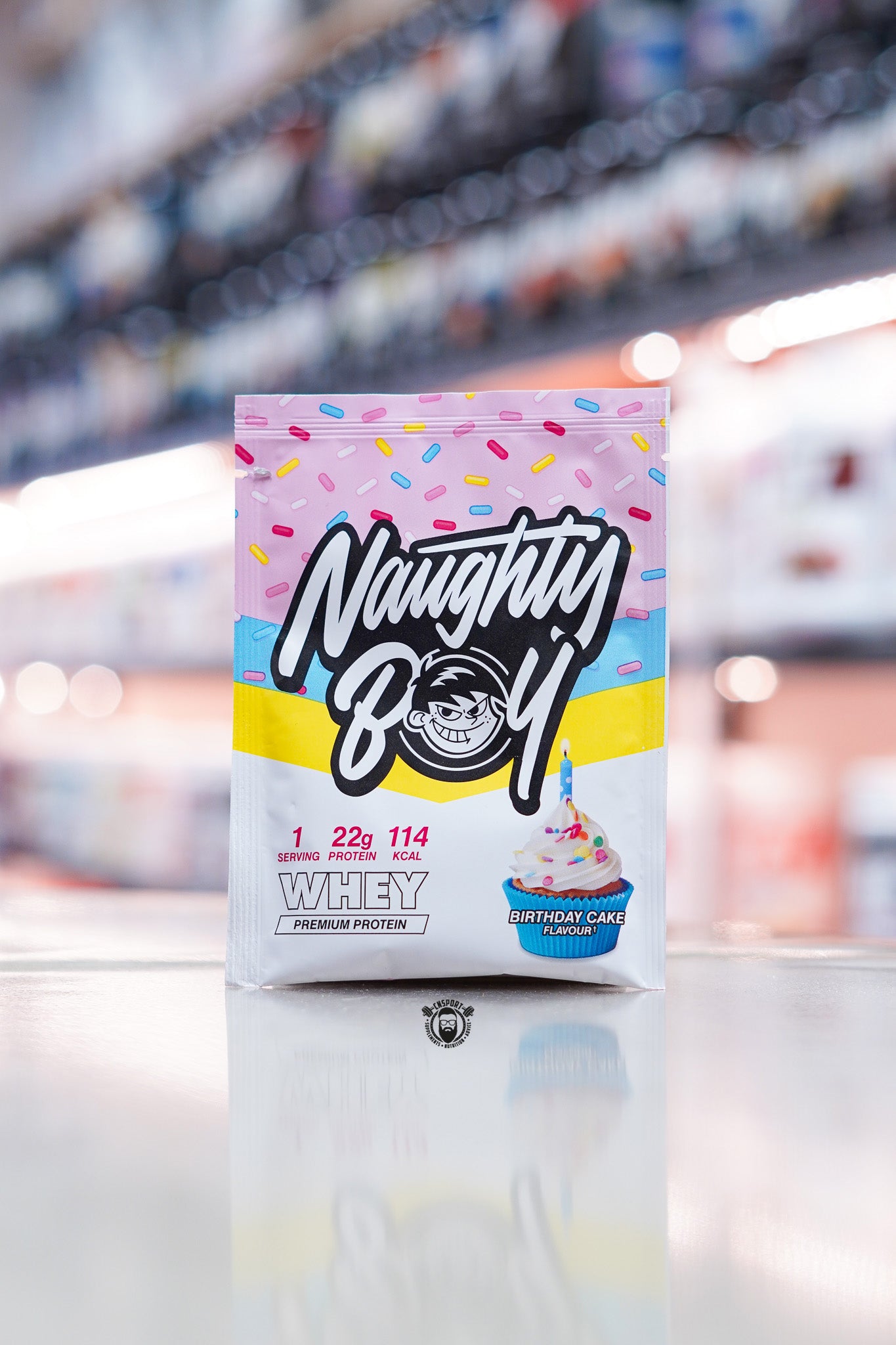 Naughty Boy Whey Protein 30g sample sachet Birthday Cake flavour on retail counter with blurred supplement store background