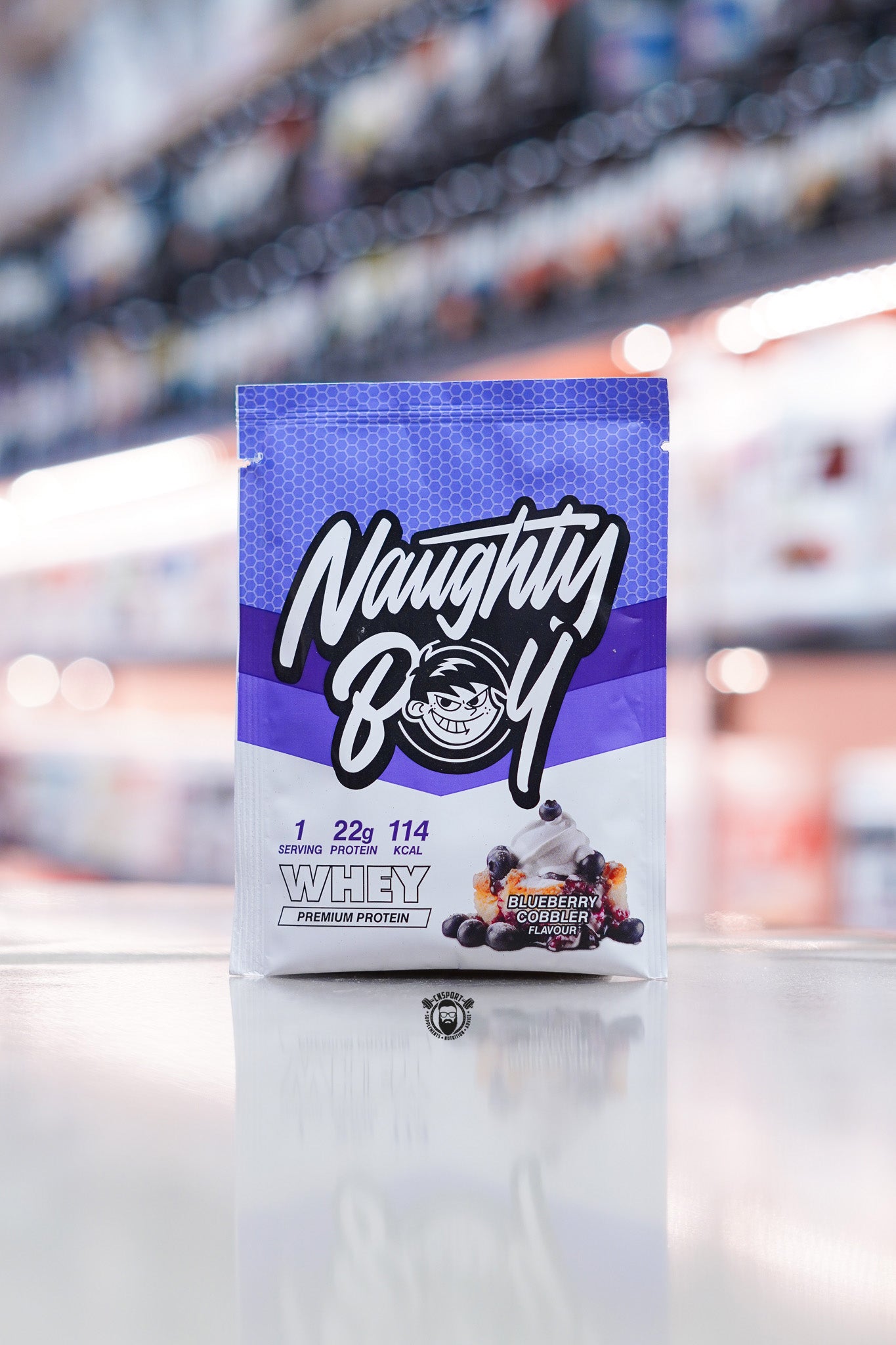 Naughty Boy Whey Protein 30g sample sachet Blueberry Cobbler flavour on retail counter with blurred supplement store background
