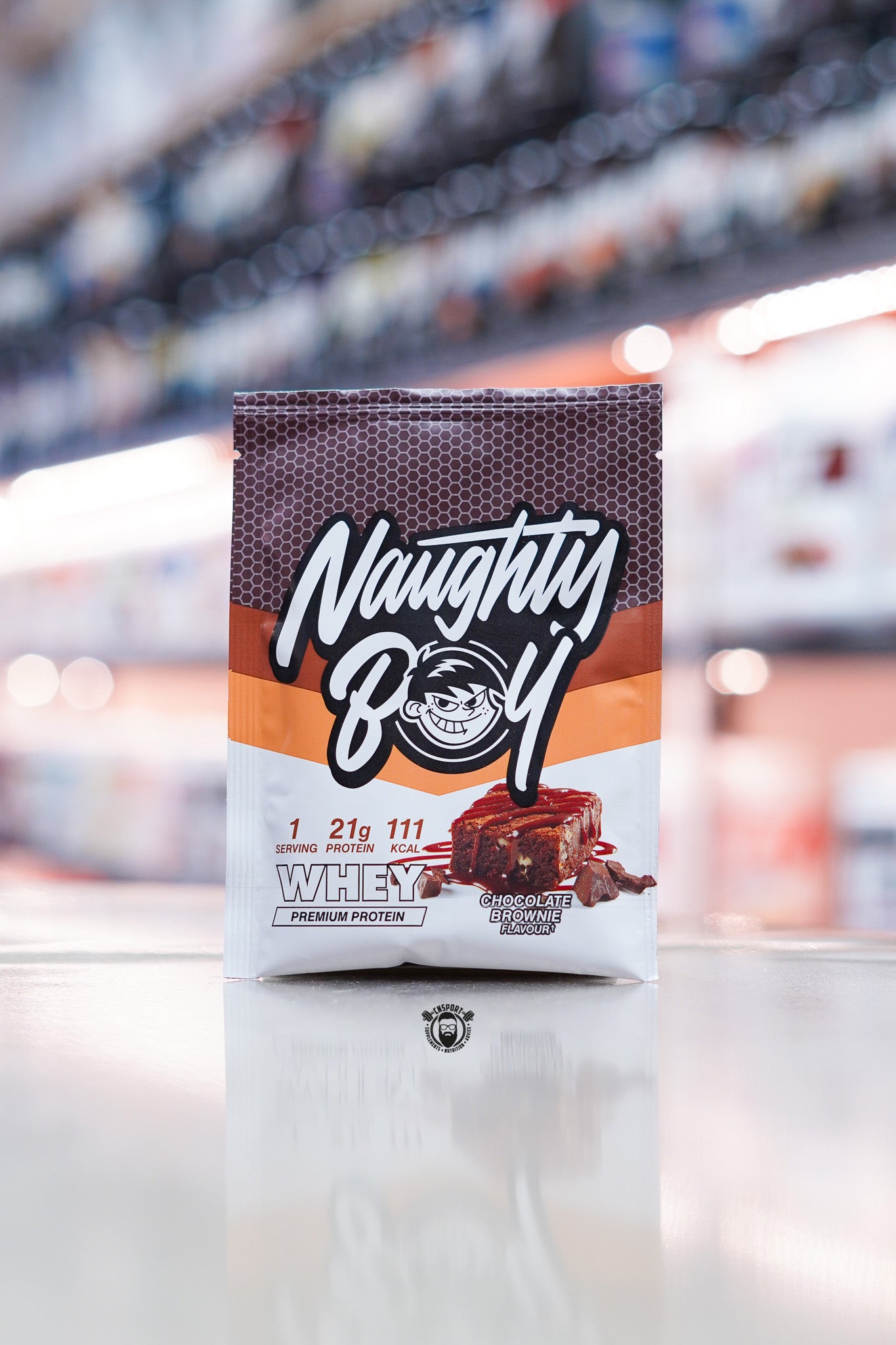 Naughty Boy Whey Protein 30g sample sachet Chocolate Brownie flavour on retail counter with blurred supplement store background