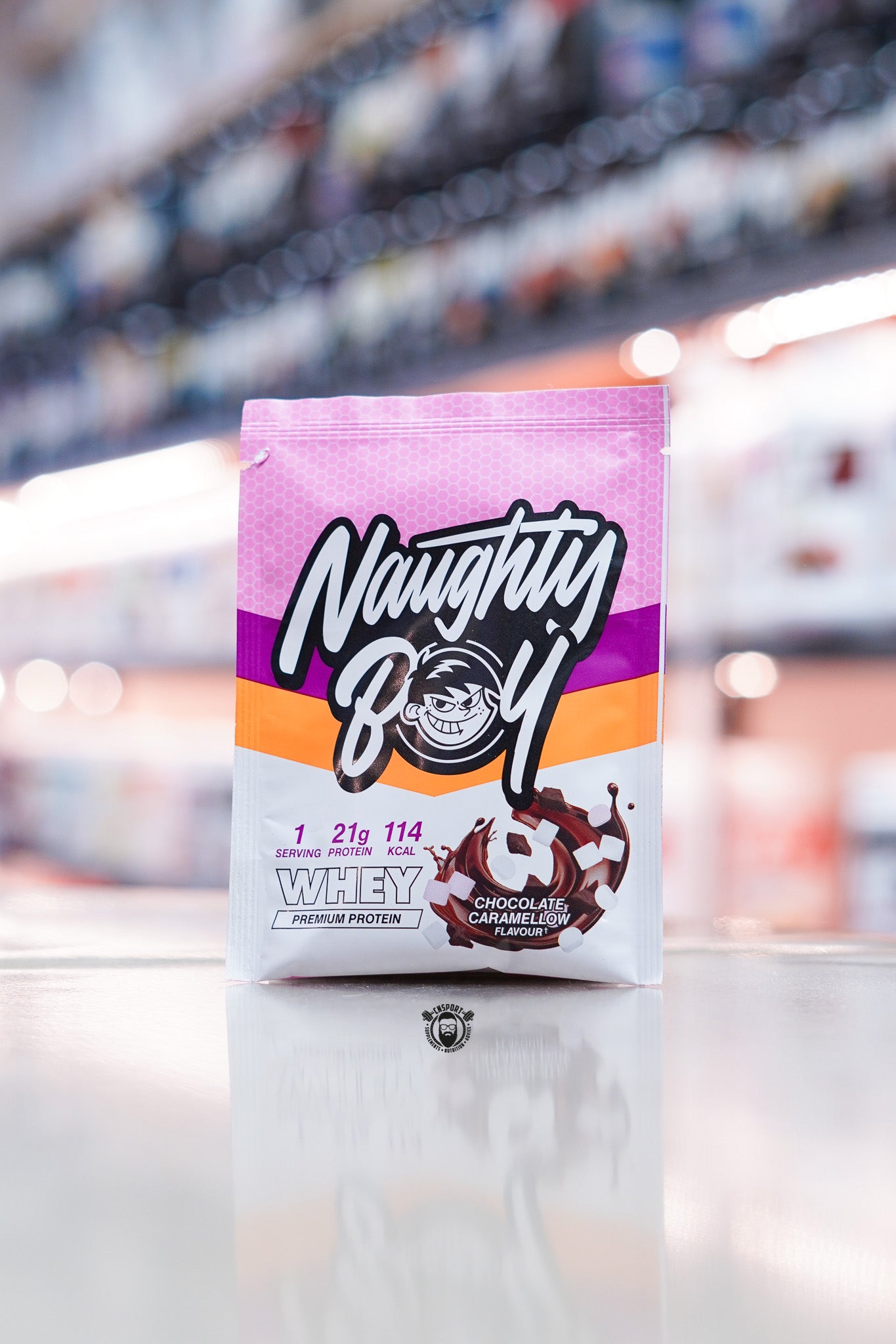 Naughty Boy Whey Protein 30g sample sachet Chocolate Caramellow flavour on retail counter with blurred supplement store background
