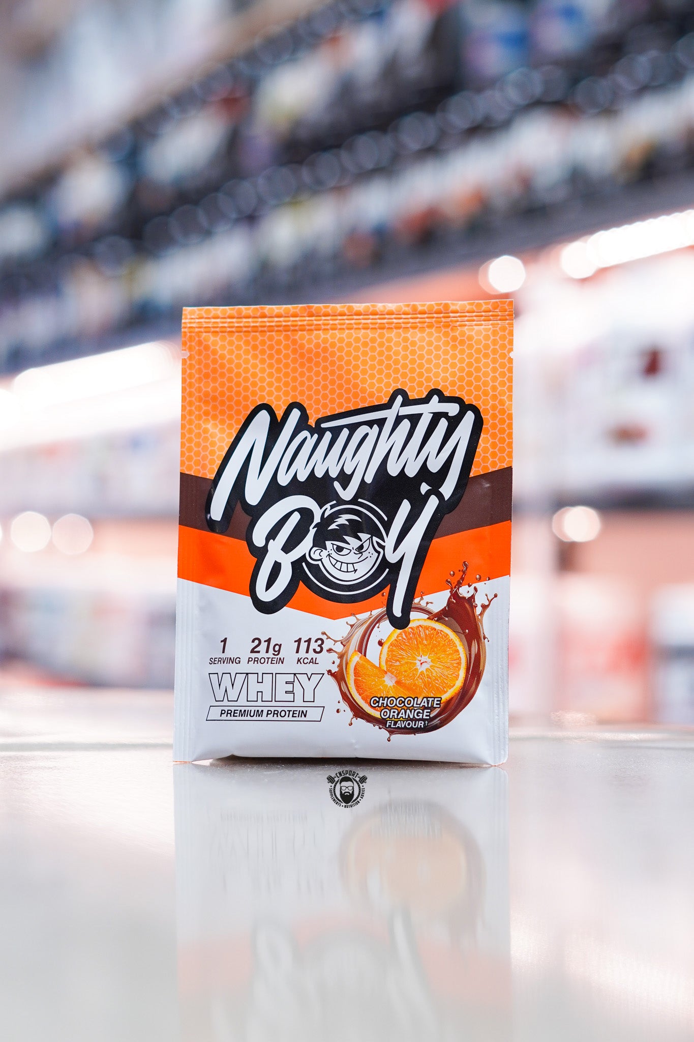 Naughty Boy Whey Protein 30g sample sachet Chocolate Orange flavour on retail counter with blurred supplement store background