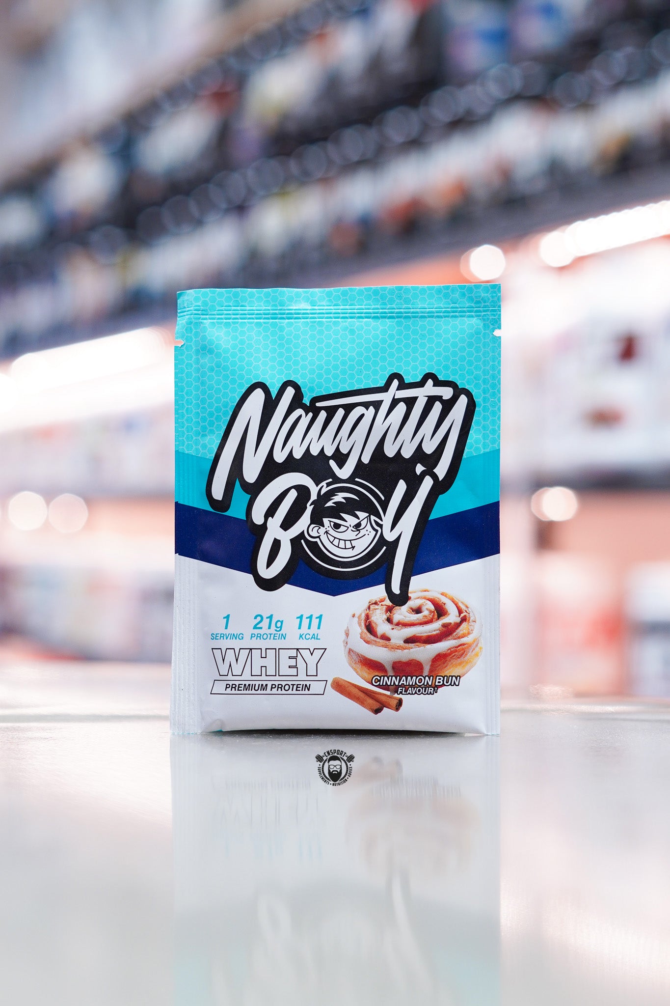 Naughty Boy Whey Protein 30g sample sachet Cinnamon Bun flavour on retail counter with blurred supplement store background