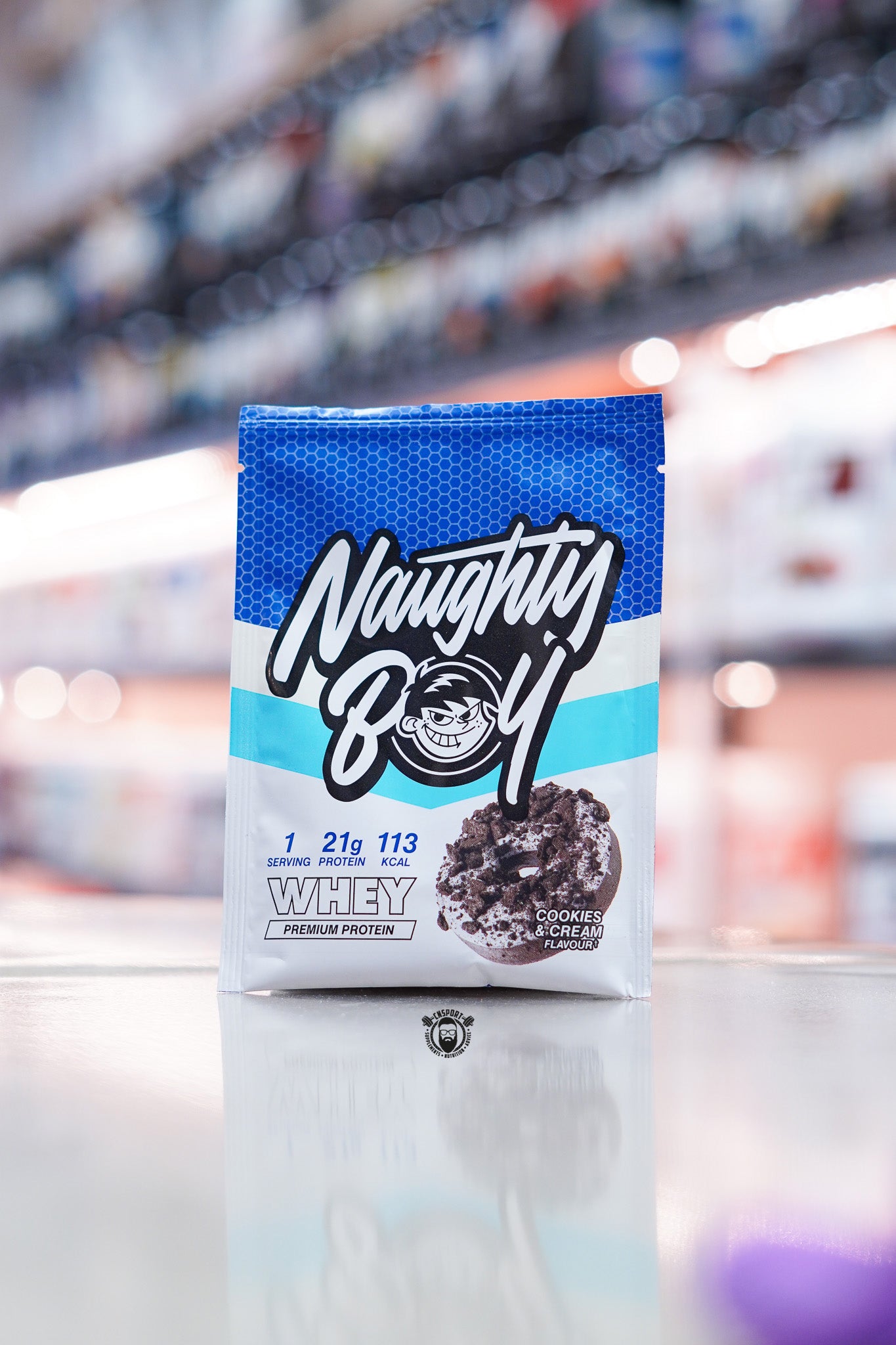 Naughty Boy Whey Protein 30g sample sachet Cookies & Cream flavour on retail counter with blurred supplement store background