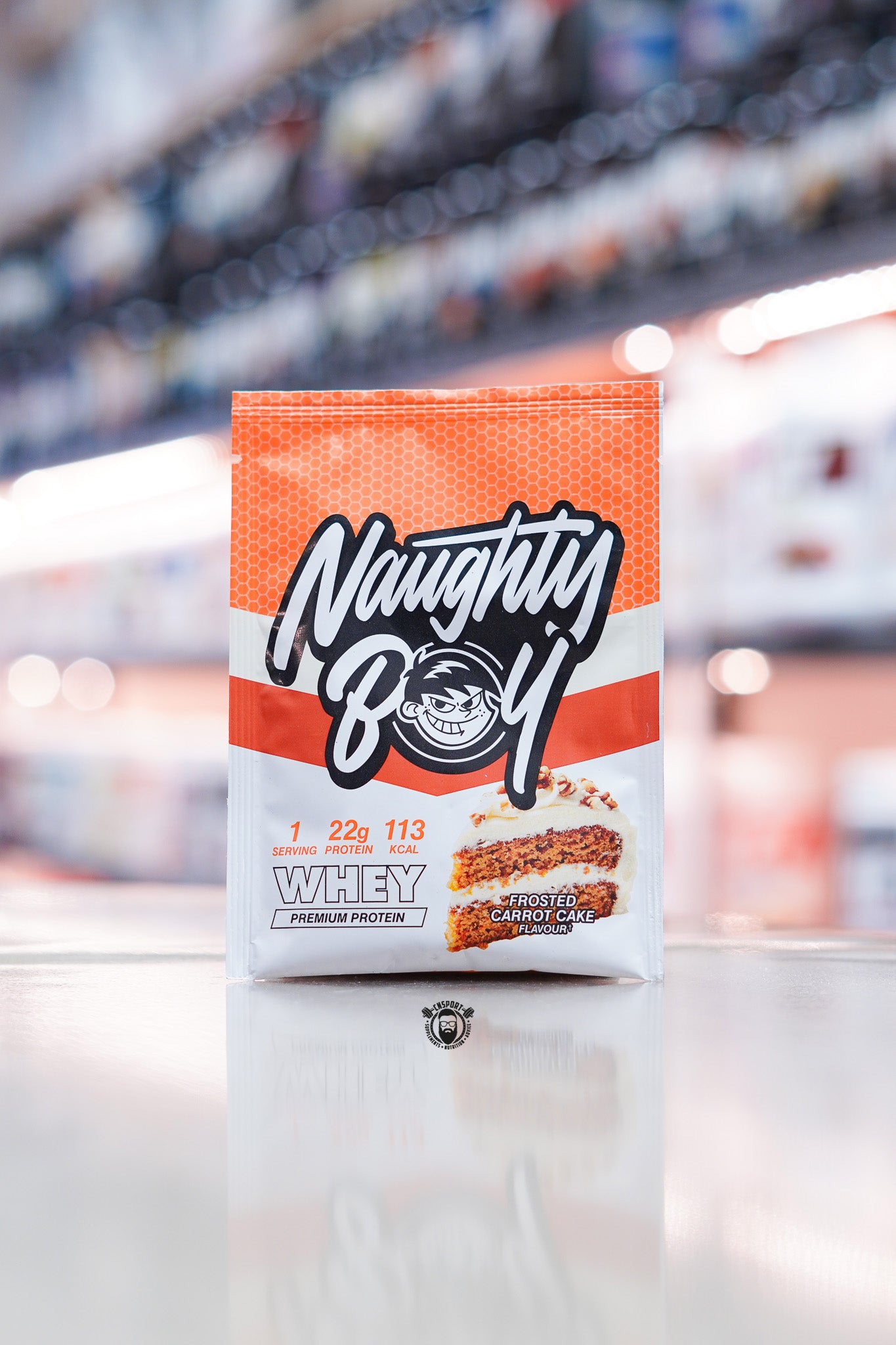 Naughty Boy Whey Protein 30g sample sachet Frosted Carrot Cake flavour on retail counter with blurred supplement store background