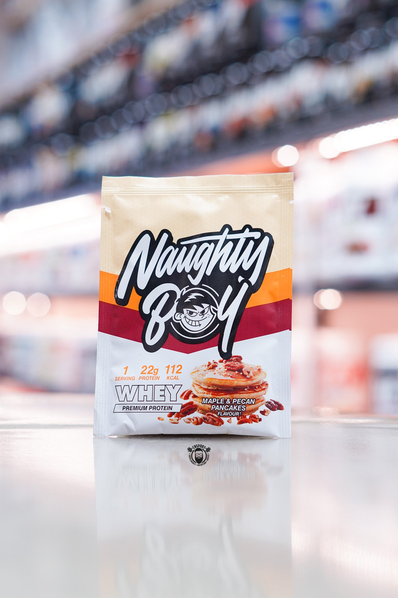 Naughty Boy Whey Protein 30g sample sachet Maple & Pecan Pancakes flavour on retail counter with blurred supplement store background
