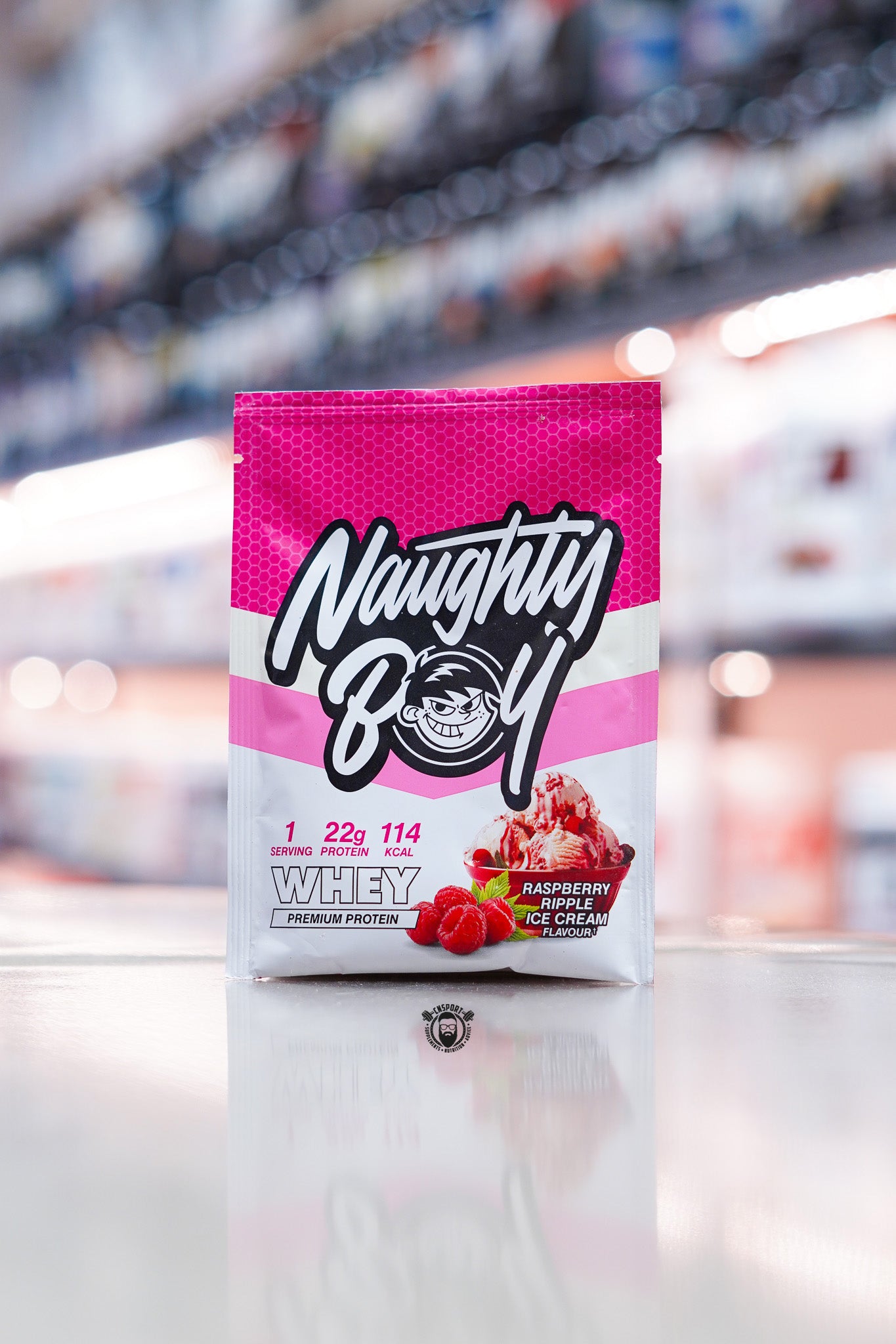 Naughty Boy Whey Protein 30g sample sachet Raspberry Ripple Ice Cream flavour on retail counter with blurred supplement store background