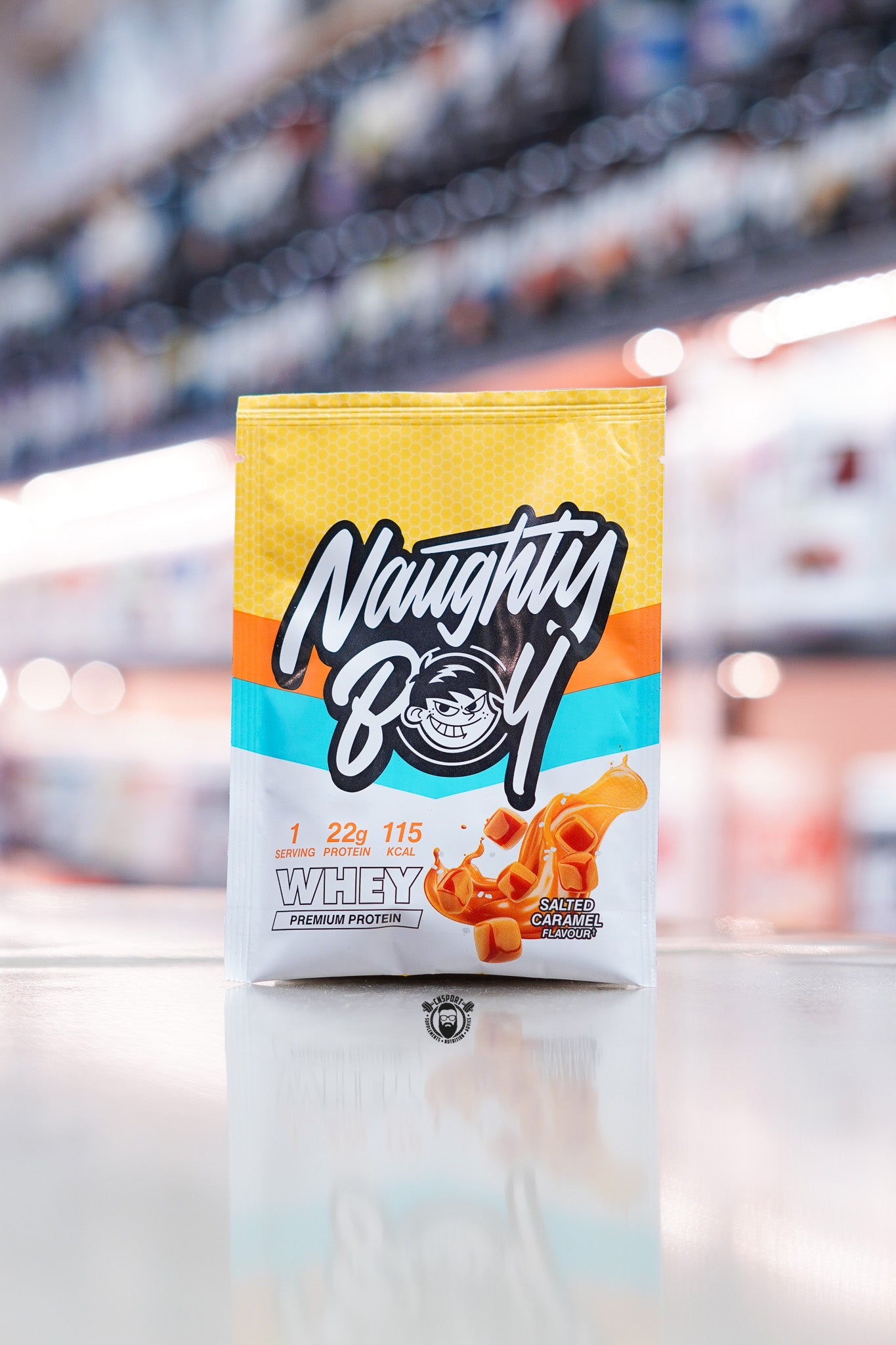 Naughty Boy Whey Protein 30g sample sachet Salted Caramel flavour on retail counter with blurred supplement store background