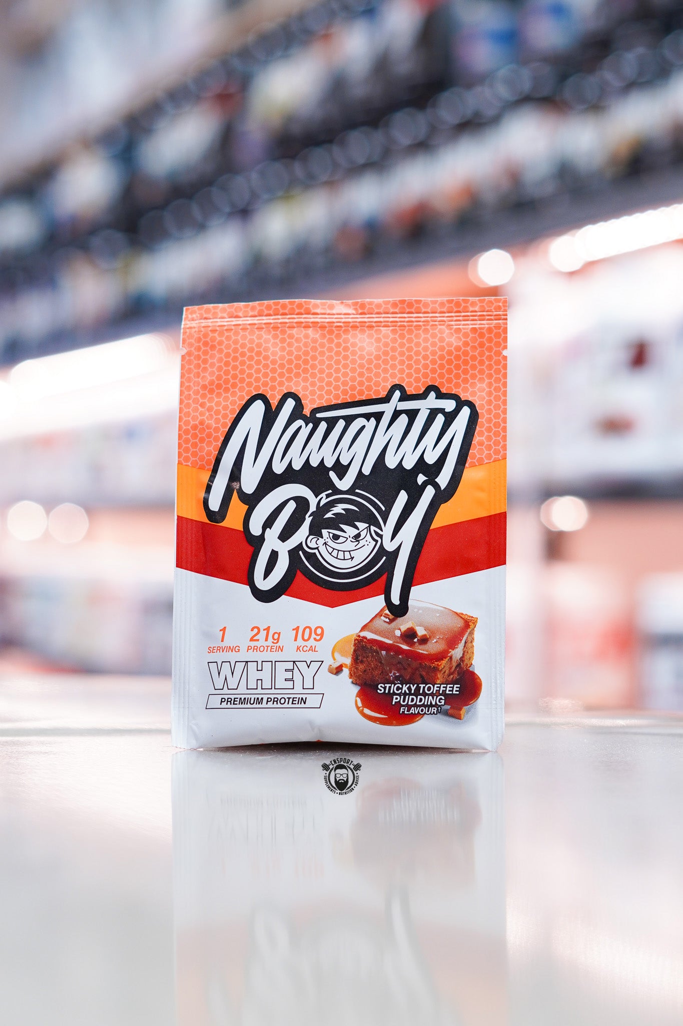 Naughty Boy Whey Protein 30g sample sachet Sticky Toffee Pudding flavour on retail counter with blurred supplement store background