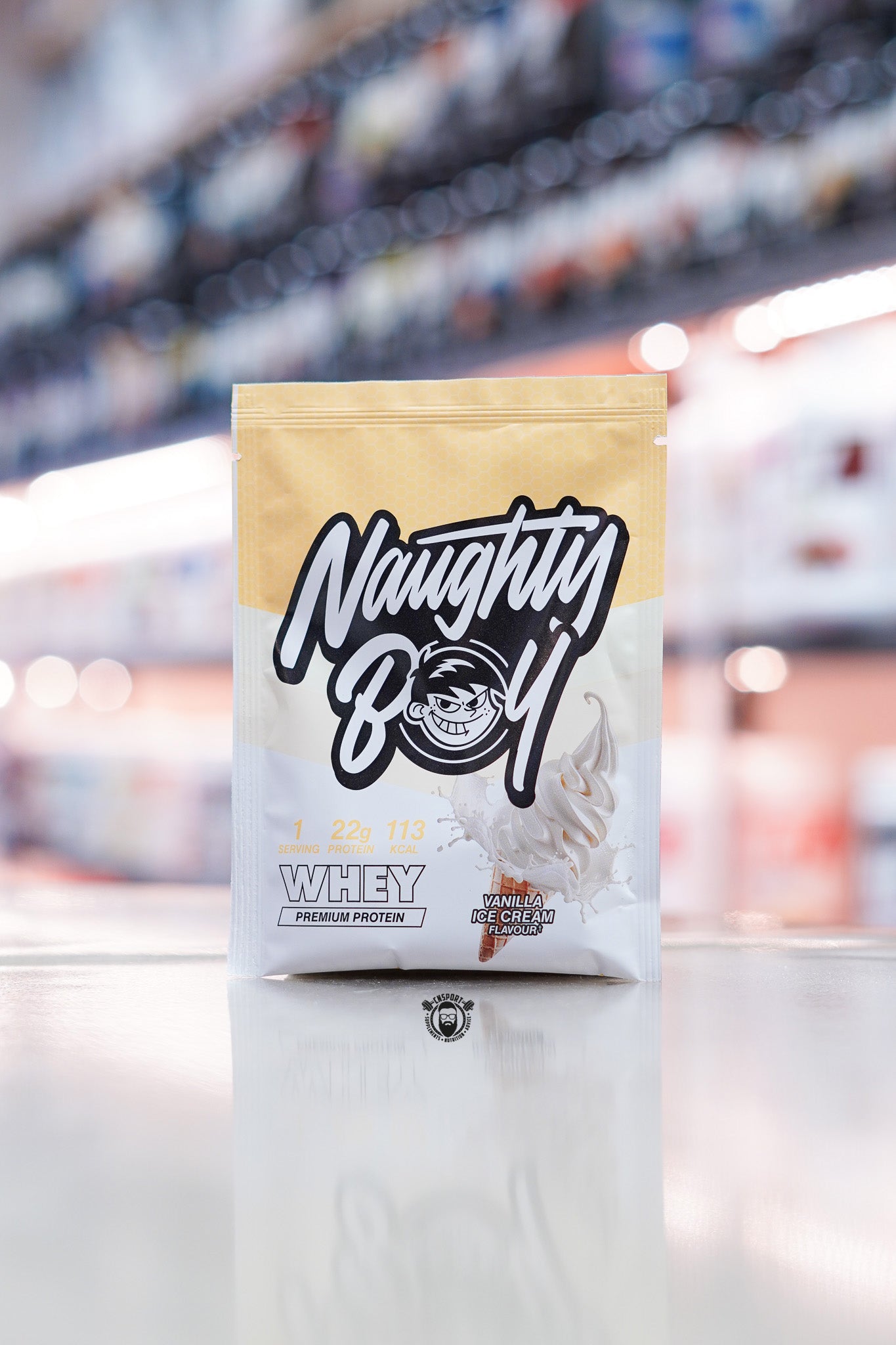 Naughty Boy Whey Protein 30g sample sachet Vanilla Ice Cream flavour on retail counter with blurred supplement store background